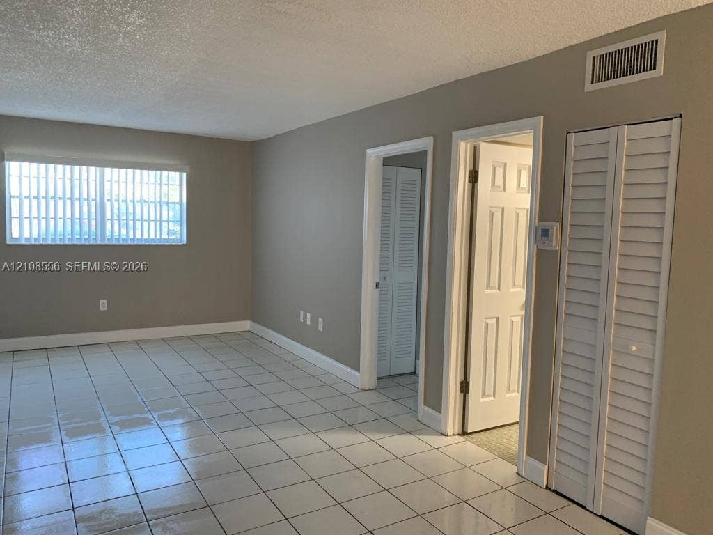 600 SW 2nd Avenue, #137, Boca Raton, FL 33432 Neutral-toned interior space featuring light-colored ceramic tile flooring, a large window with vertical blinds, and dual white louvered closet doors