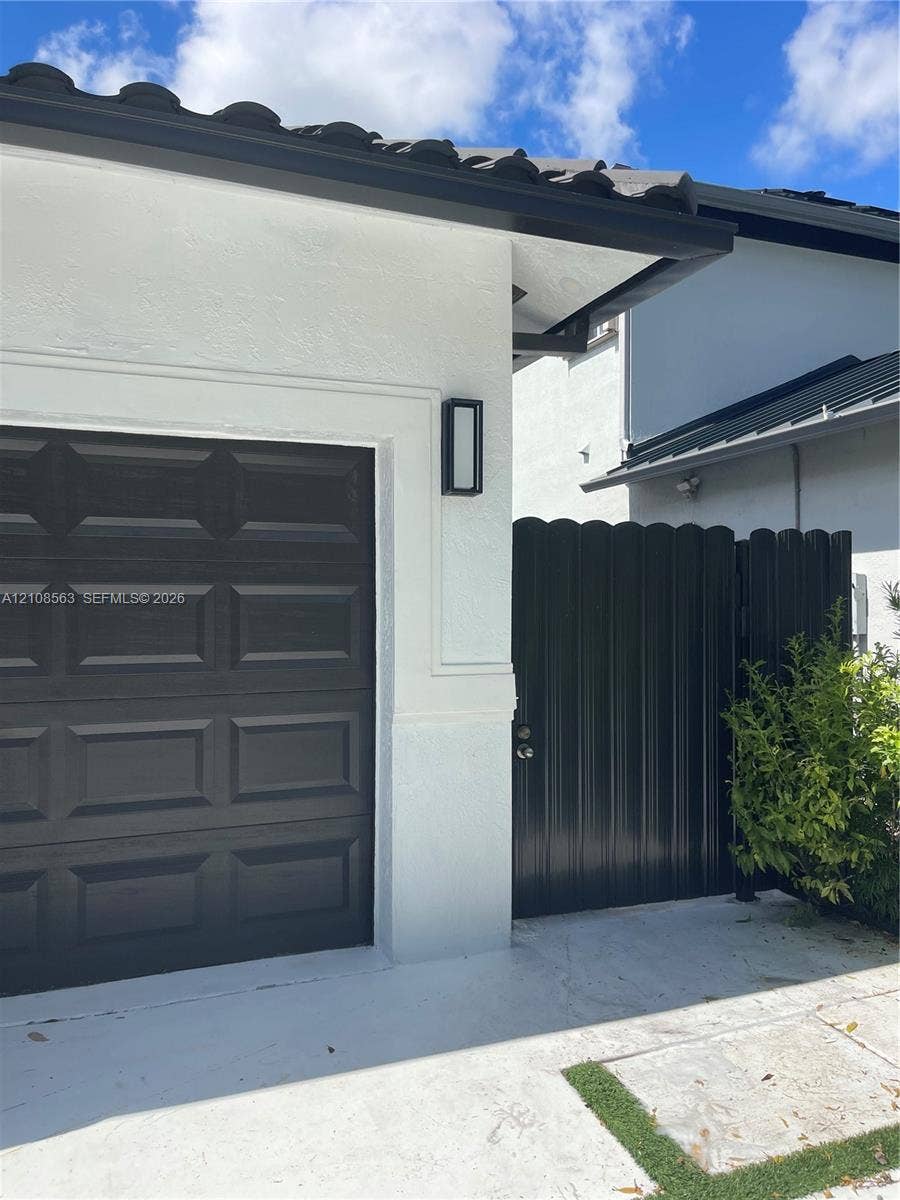 14554 SW 161 Court, #Studio, Miami, Miami, FL 33196 Stucco exterior featuring a dark-finish paneled garage door, scalloped privacy gate, and modern rectangular wall-mounted light fixture under a tiled roofline