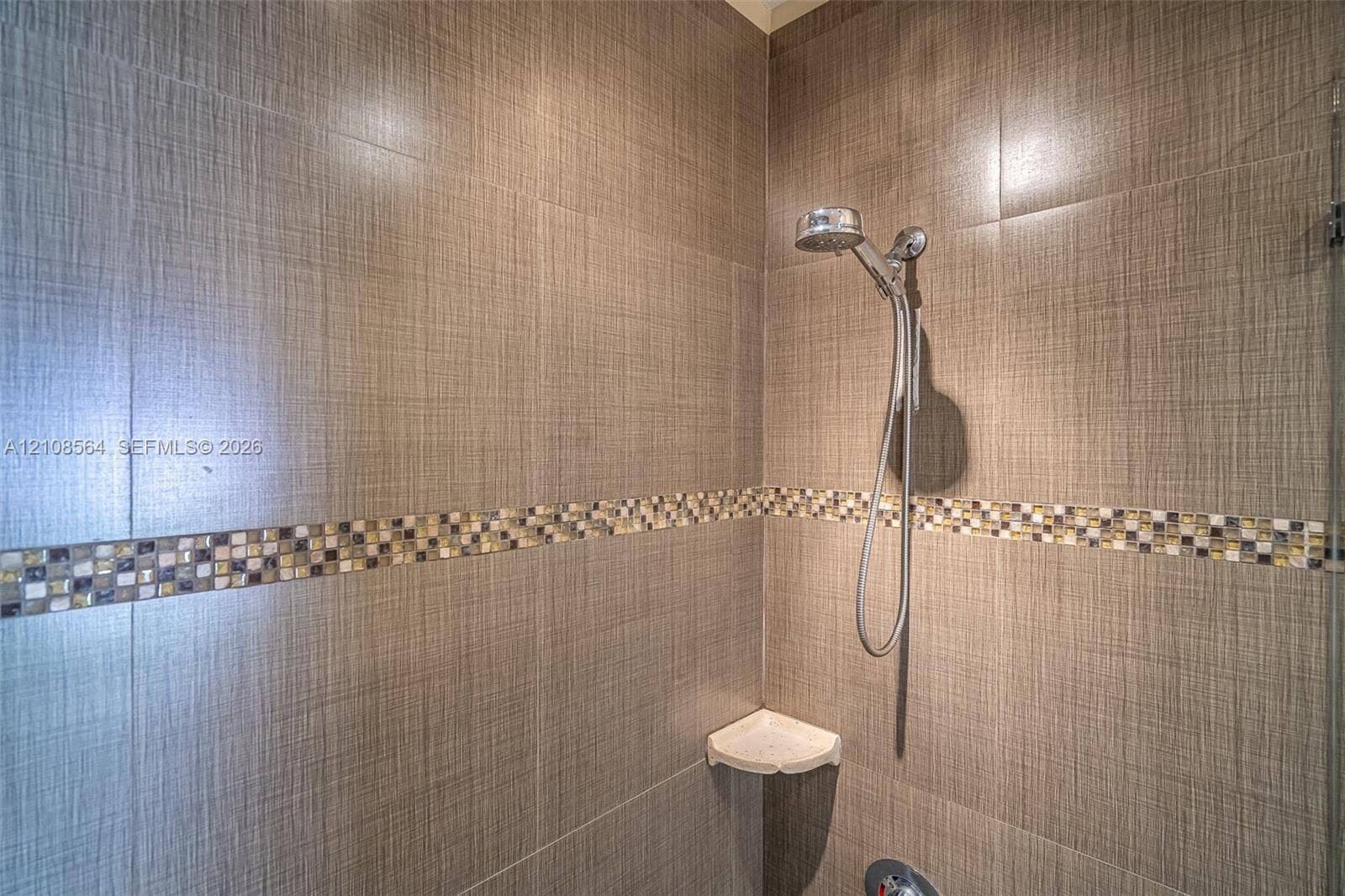 3111 NW 86th Avenue, Sunrise, FL 33351 Shower enclosure featuring large-format textured wall tile, decorative mosaic tile inlay, chrome handheld shower fixture, and a built-in corner shelf
