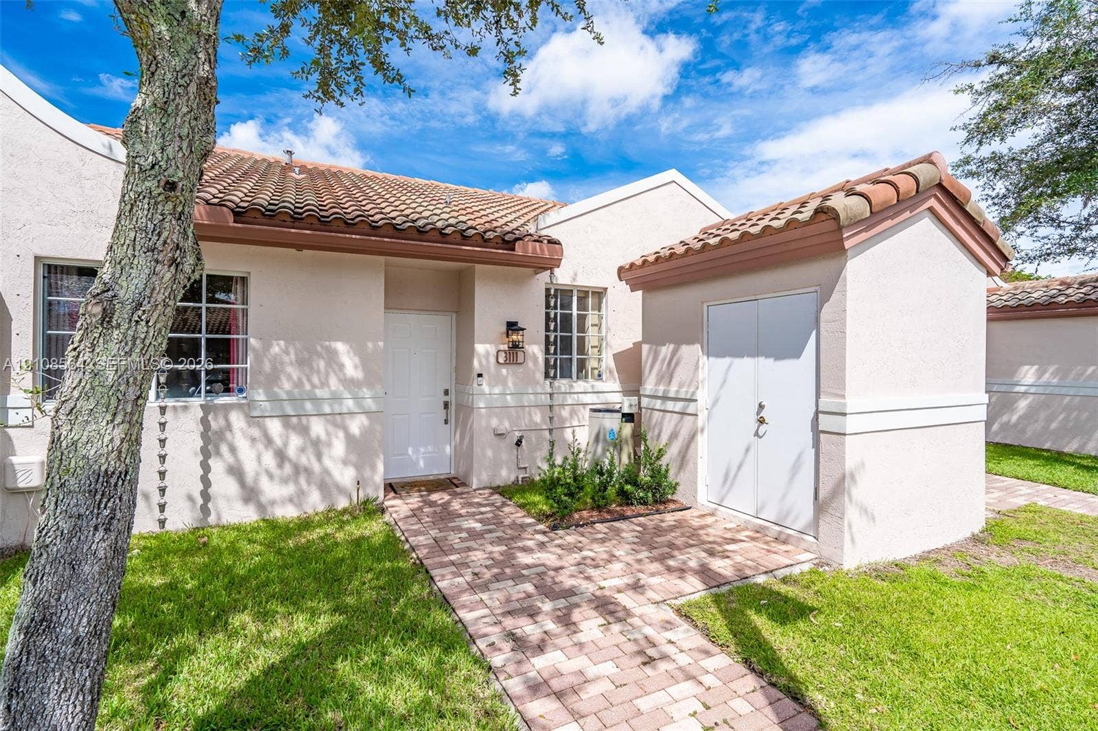 3111 NW 86th Avenue, Sunrise, FL 33351 Stucco exterior featuring a clay tile roof, brick paver walkway, white paneled front door, and an attached exterior storage structure