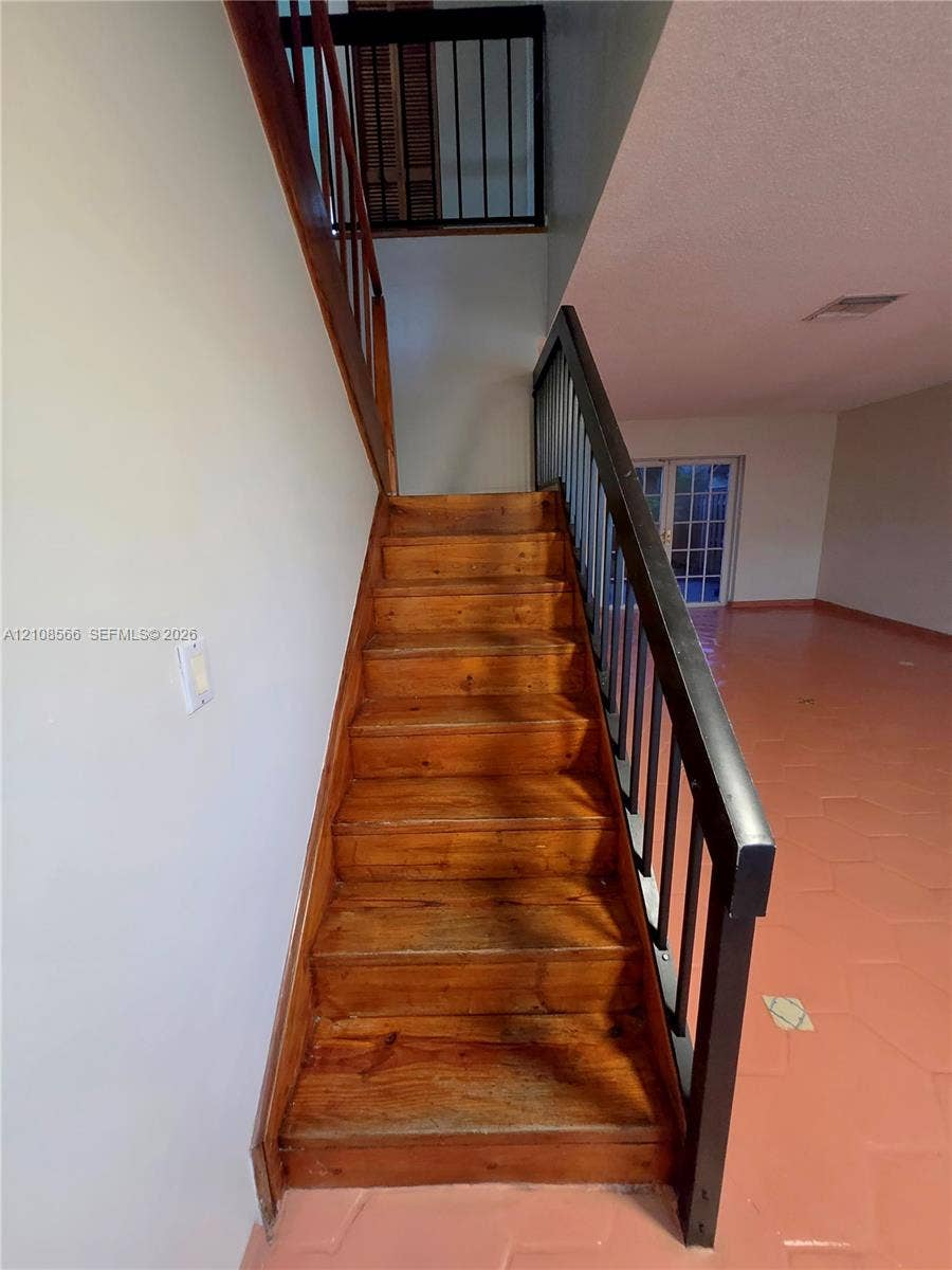 Staircase featuring natural wood treads, a wooden handrail, and dark metal balusters, complemented by tile flooring