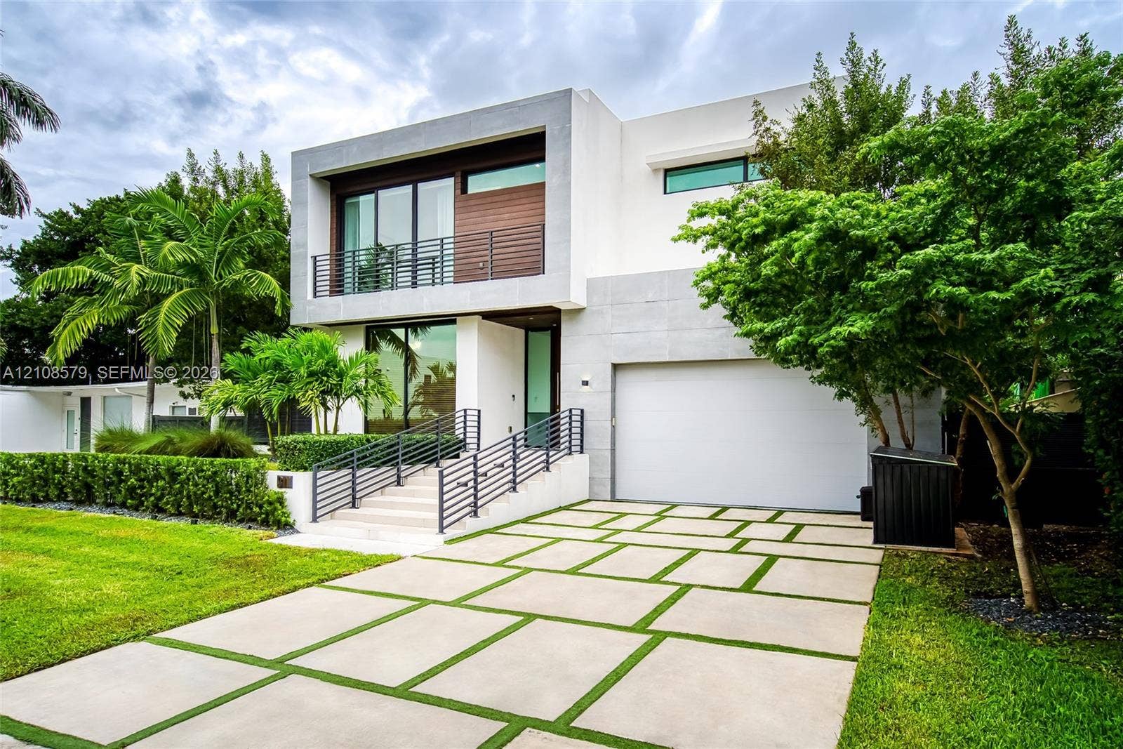 311 189th Street, #0, Sunny Isles Beach, Sunny Isles Beach, FL 33160 Modern two-story architectural exterior featuring smooth-finish stucco walls, natural wood-paneled accents, a linear upper-level balcony with metal railings, and a geometric concrete paver driveway with integrated turf strips