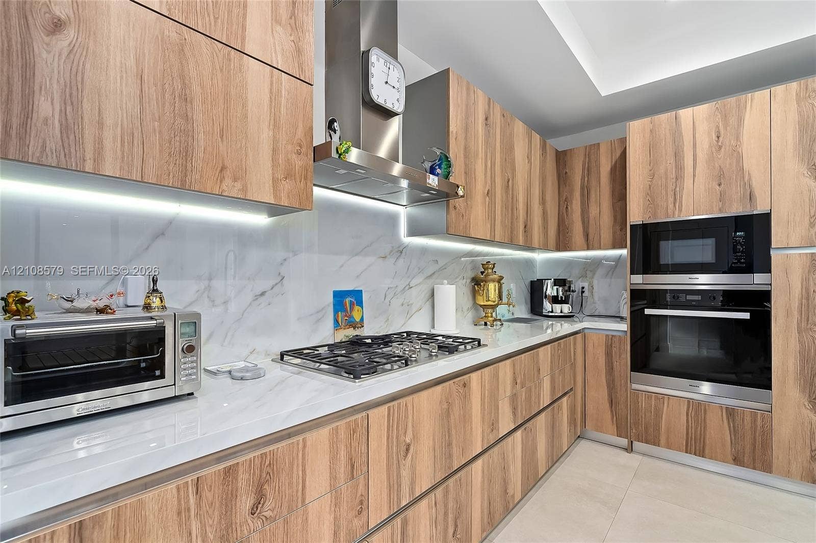 Kitchen featuring wood-finish cabinetry, stone-surface countertops, integrated under-cabinet lighting, a stainless steel gas cooktop with chimney-style range hood, and a built-in stainless steel oven and microwave combination