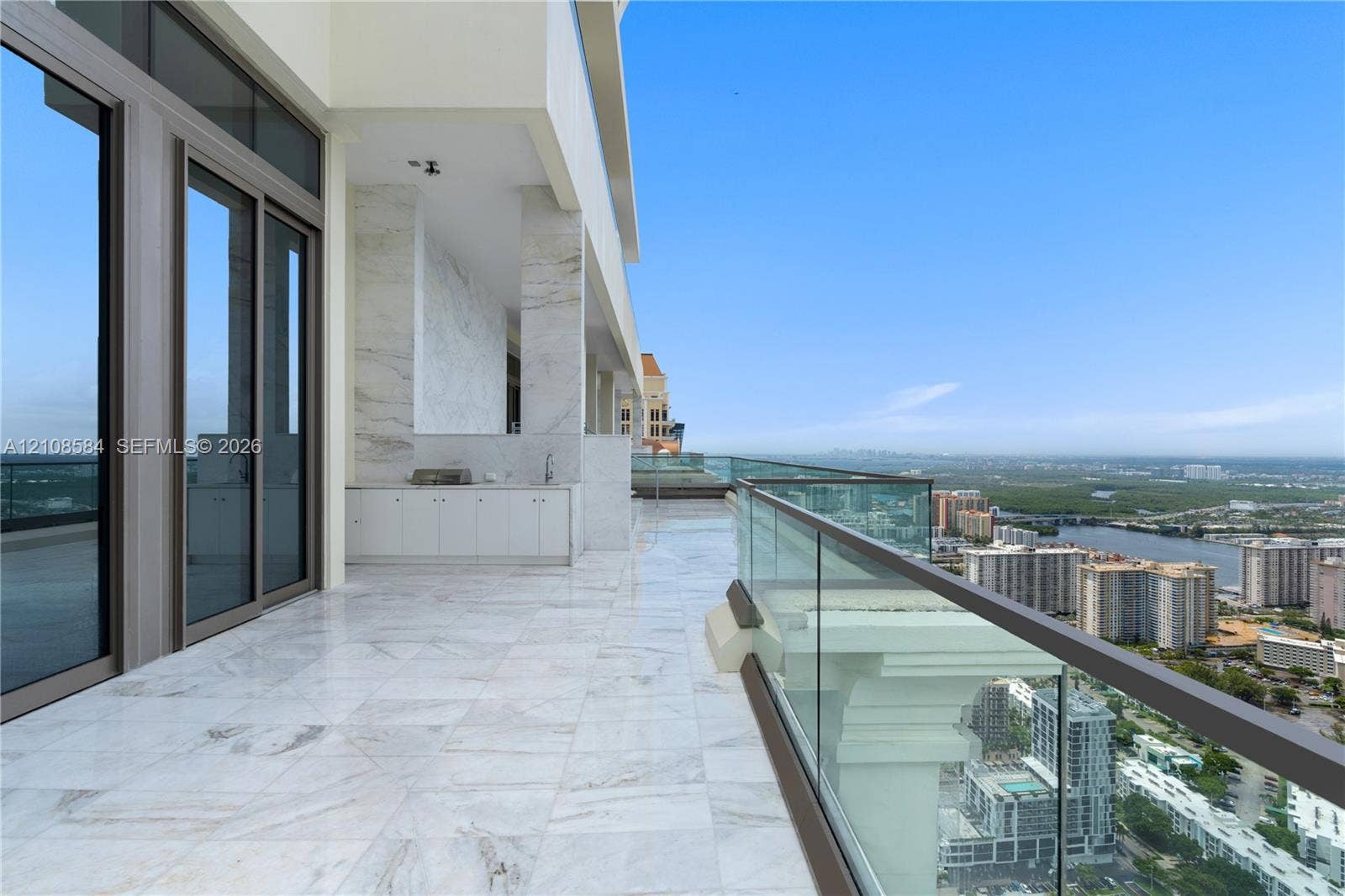 Expansive covered balcony featuring stone flooring, integrated outdoor cabinetry with a built-in grill, stone-clad structural columns, and a frameless glass railing