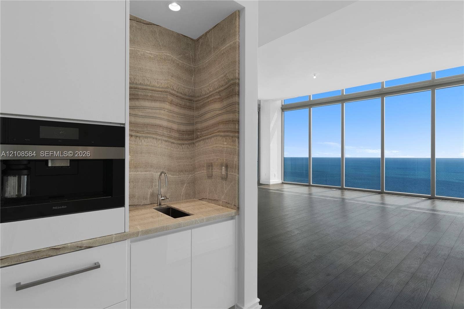Wet bar featuring built-in coffee system, stone slab backsplash, integrated sink, and white cabinetry, set against floor-to-ceiling windows with ocean scenery and dark wood-finish flooring