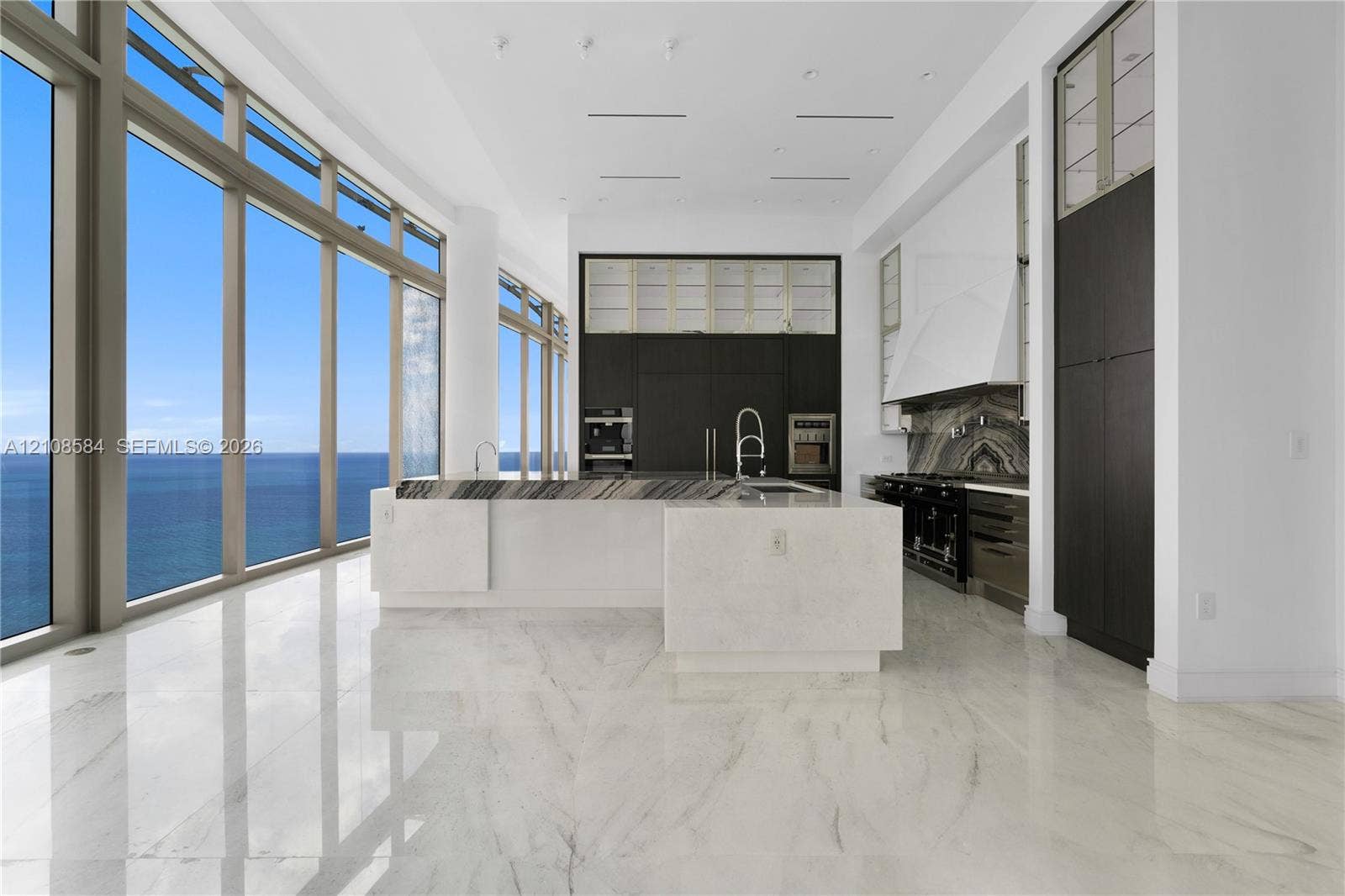 Floor-to-ceiling windows with ocean scenery, expansive center island with stone countertop, custom floor-to-ceiling dark cabinetry, high-gloss stone flooring, and professional-grade range with decorative stone backsplash