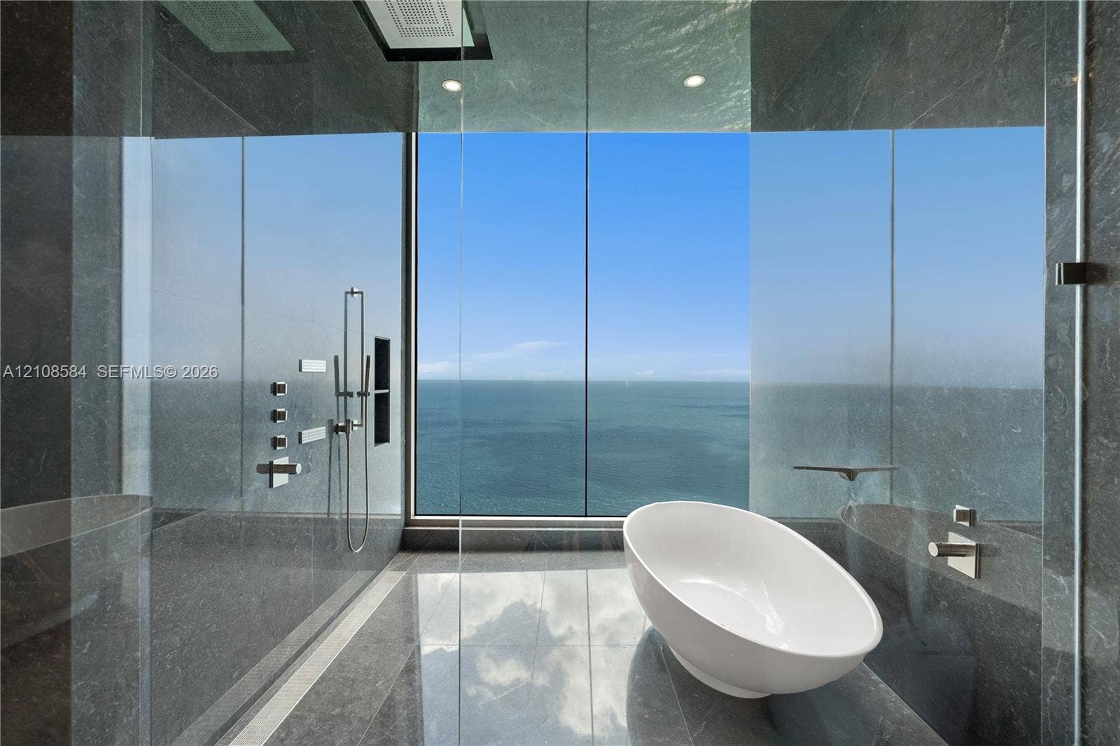 Full-height stone wall tiling, glass-enclosed shower featuring a rainfall showerhead and wall-mounted controls, floor-to-ceiling windows, and a freestanding oval soaking tub