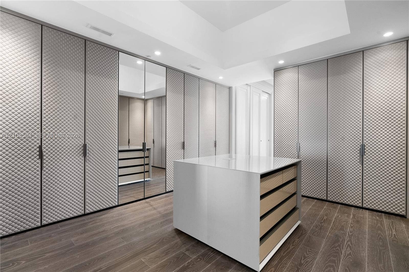 Floor-to-ceiling built-in cabinetry with diamond-quilted paneling, full-height mirrored wardrobe door, central island with smooth countertop, recessed lighting, and wood-finish flooring