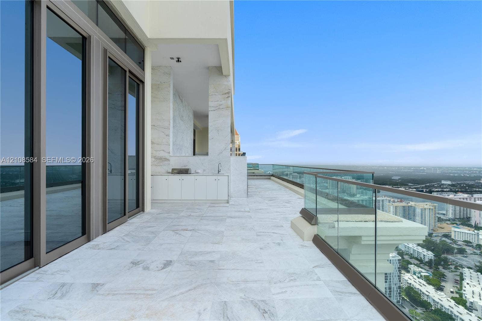 Expansive covered terrace featuring large-format stone tile flooring, built-in outdoor cabinetry with a sink, stone-clad structural columns, and a glass-paneled railing
