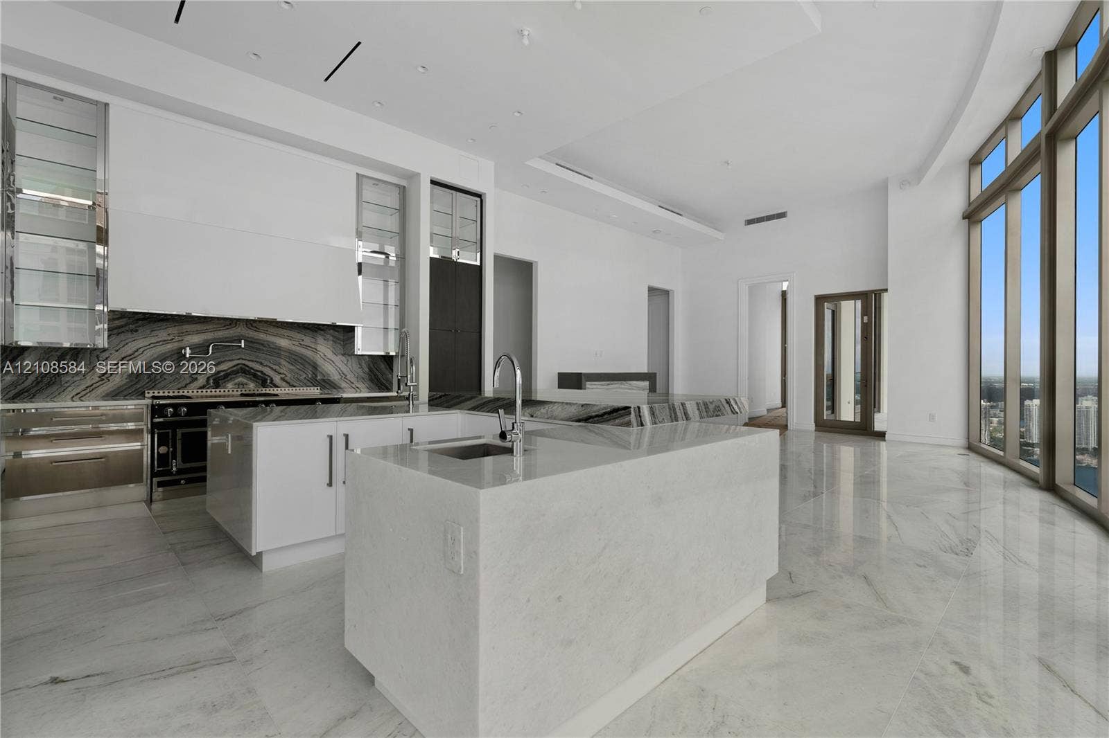 Contemporary kitchen featuring floor-to-ceiling windows, high-gloss cabinetry, stone-surfaced island with integrated sink, stone backsplash, and polished stone flooring