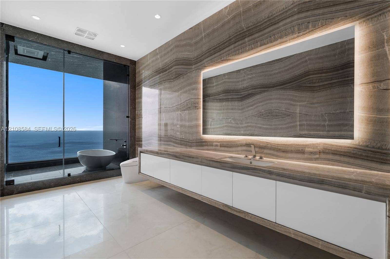 Full-height natural stone wall cladding, floating white cabinetry with integrated countertop, recessed mirror with perimeter lighting, large-format polished flooring, and floor-to-ceiling glass panel enclosing a soaking tub
