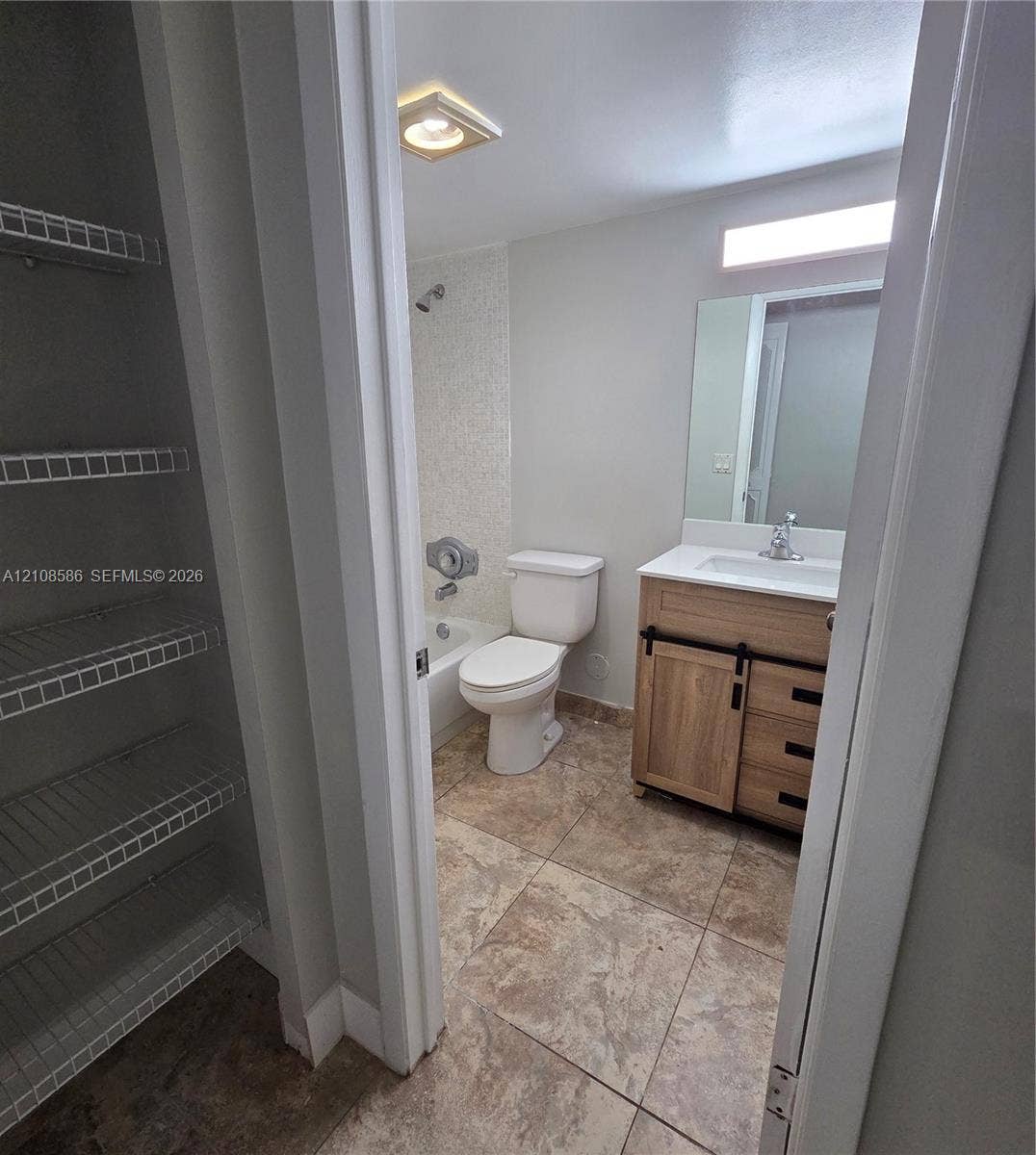 6075 N Sabal Palm Boulevard, #110, Tamarac, FL 33319 Bathroom featuring a wood-finish vanity with a white countertop, large wall-mounted mirror, tile flooring, bathtub with a tiled shower surround, and a walk-in storage area with wire shelving