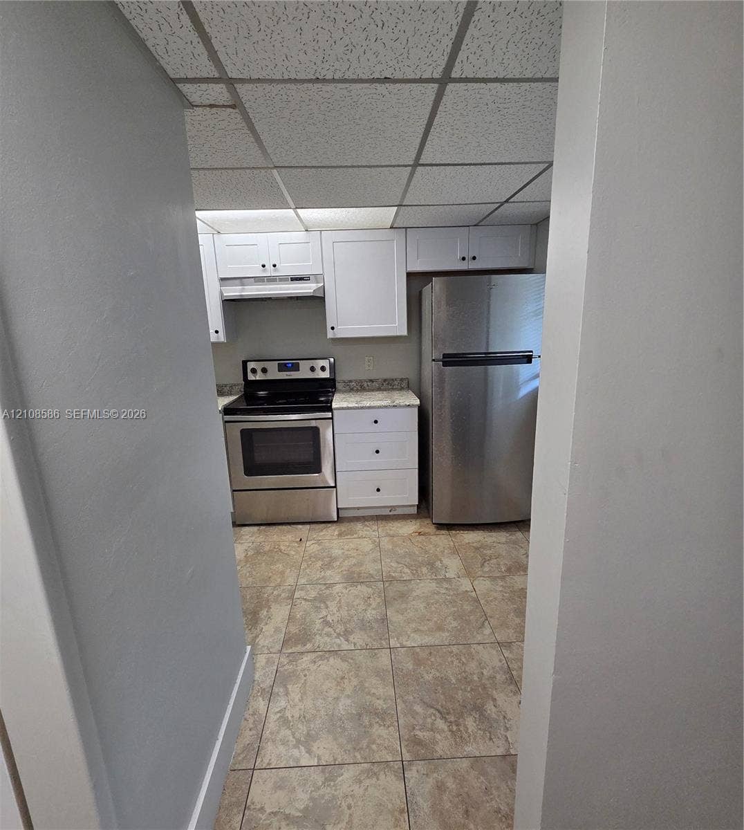 6075 N Sabal Palm Boulevard, #110, Tamarac, FL 33319 Kitchen featuring white shaker-style cabinetry, stainless steel appliances, speckled stone-finish countertops, tile flooring, and drop ceiling panels