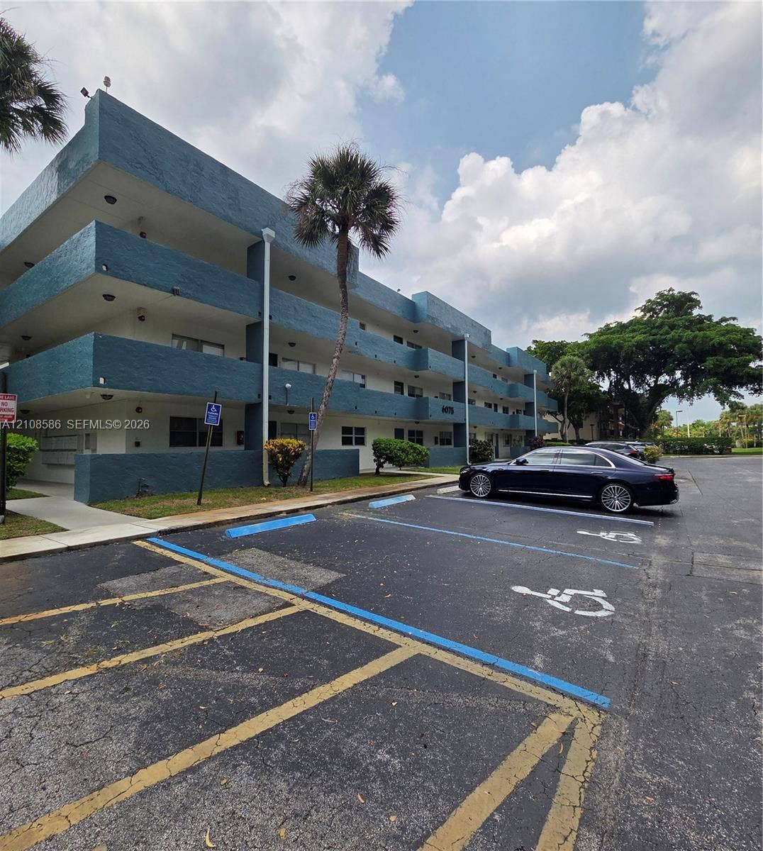 6075 N Sabal Palm Boulevard, #110, Tamarac, FL 33319 Three-story multi-unit structure featuring a textured blue facade, recessed balconies, and paved surface parking