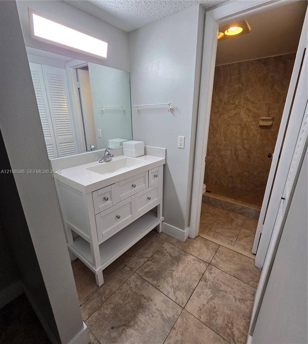 6075 N Sabal Palm Boulevard, #110, Tamarac, FL 33319 White vanity with integrated countertop and sink, large rectangular wall mirror, overhead linear lighting, and tile flooring with matching wall-tiled shower enclosure