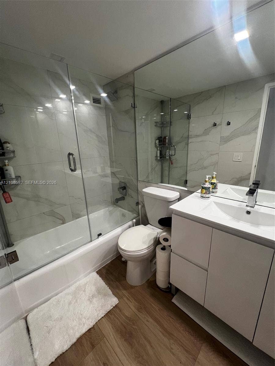 10185 Collins Avenue, #515, Bal Harbour, Bal Harbour, FL 33154 Bathroom featuring a vanity with a white countertop and rectangular basin, full-width wall-mounted mirror, wood-finish flooring, and a glass-enclosed shower with large-format wall tiling