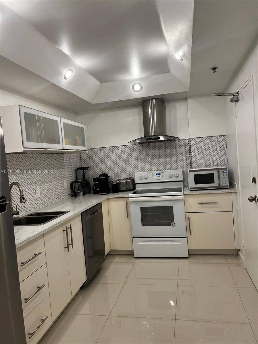 10185 Collins Avenue, #515, Bal Harbour, Bal Harbour, FL 33154 Kitchen featuring high-gloss cabinetry, stone-finish countertops, herringbone-patterned tile backsplash, stainless steel chimney-style range hood, and recessed tray ceiling lighting