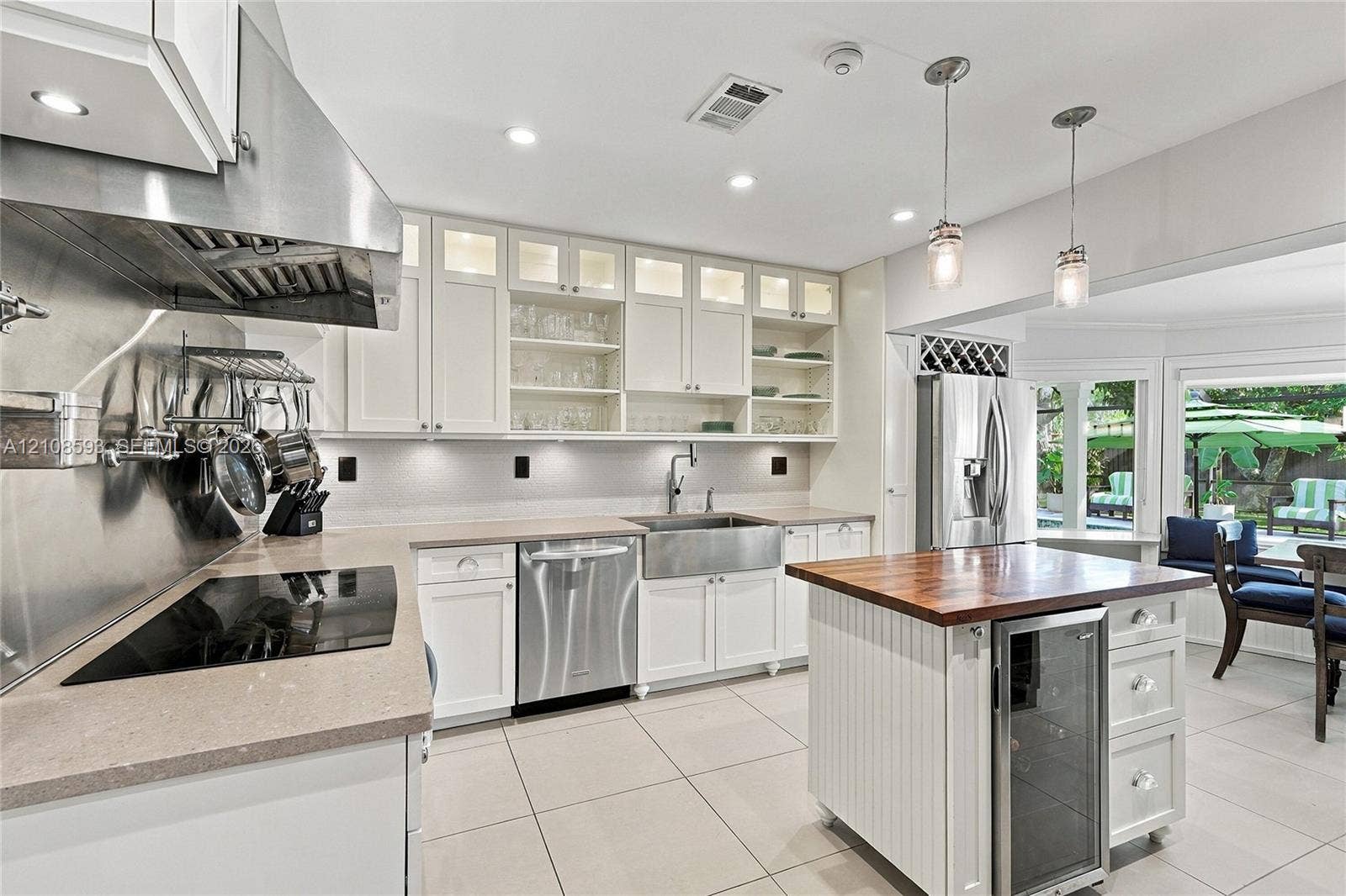 8230 SW 151st Street, Palmetto Bay, FL 33158 Kitchen featuring white cabinetry with glass-front uppers, stainless steel farmhouse sink, professional-grade range hood, butcher-block topped island with built-in wine refrigeration, and stone-finish countertops