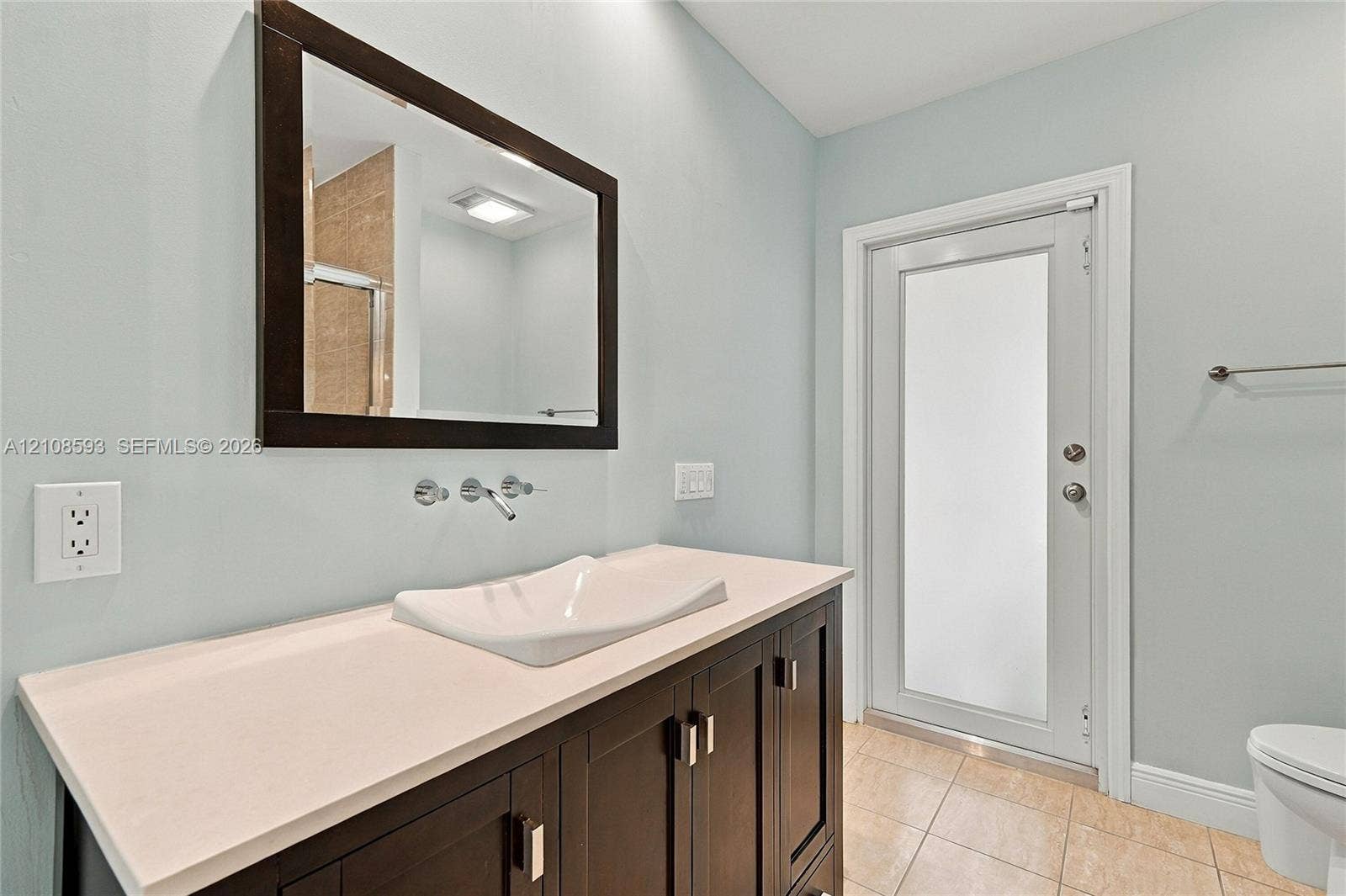 8230 SW 151st Street, Palmetto Bay, FL 33158 Vanity featuring a stone-surface countertop, rectangular vessel sink, wall-mounted faucet, framed wall mirror, and dark-finish cabinetry with metallic hardware, paired with a door featuring a frosted glass insert