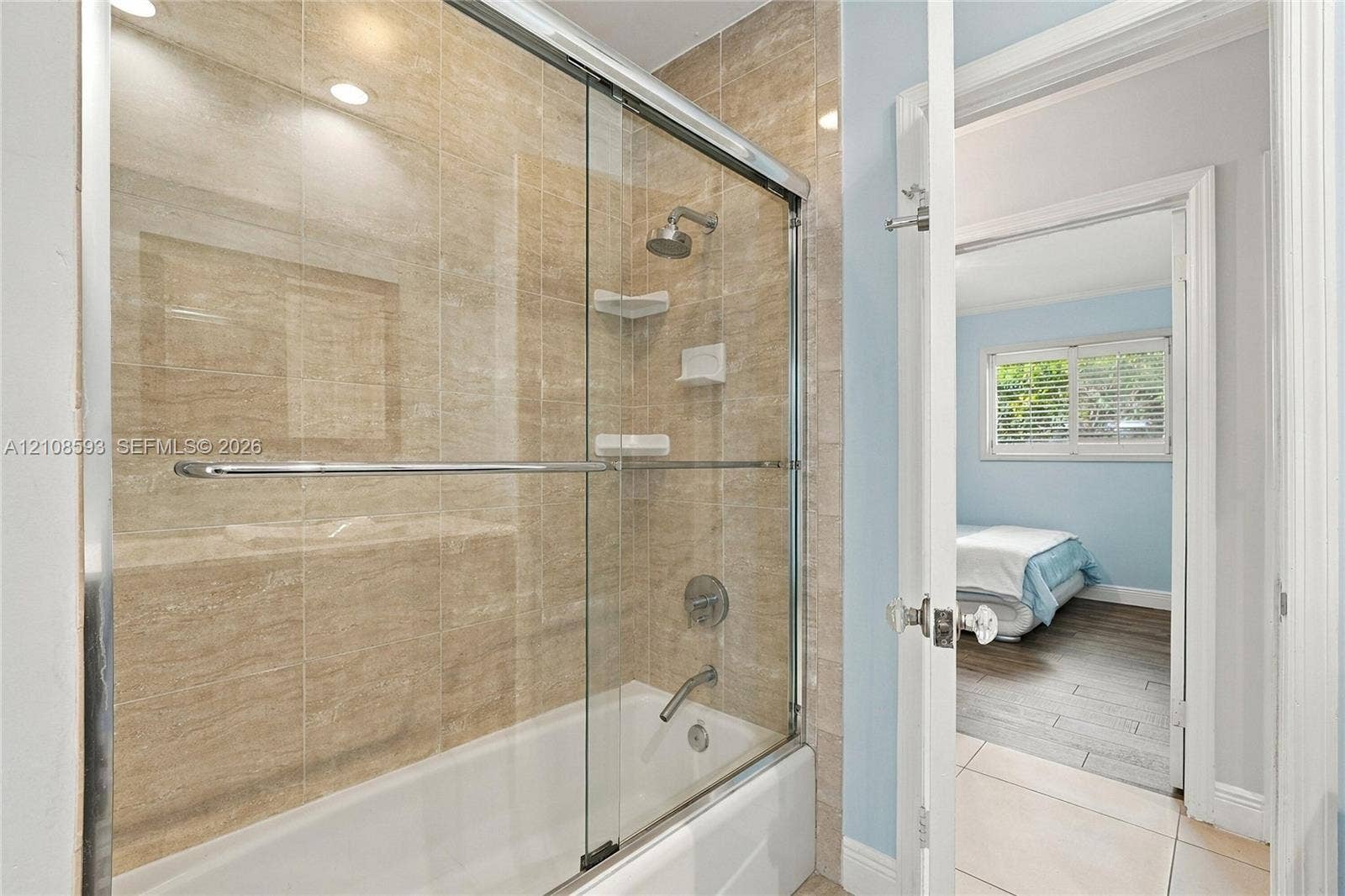 8230 SW 151st Street, Palmetto Bay, FL 33158 Bathtub and shower combination with floor-to-ceiling neutral tone tile surround, sliding glass enclosure, integrated corner shelving, and chrome faucet fixtures