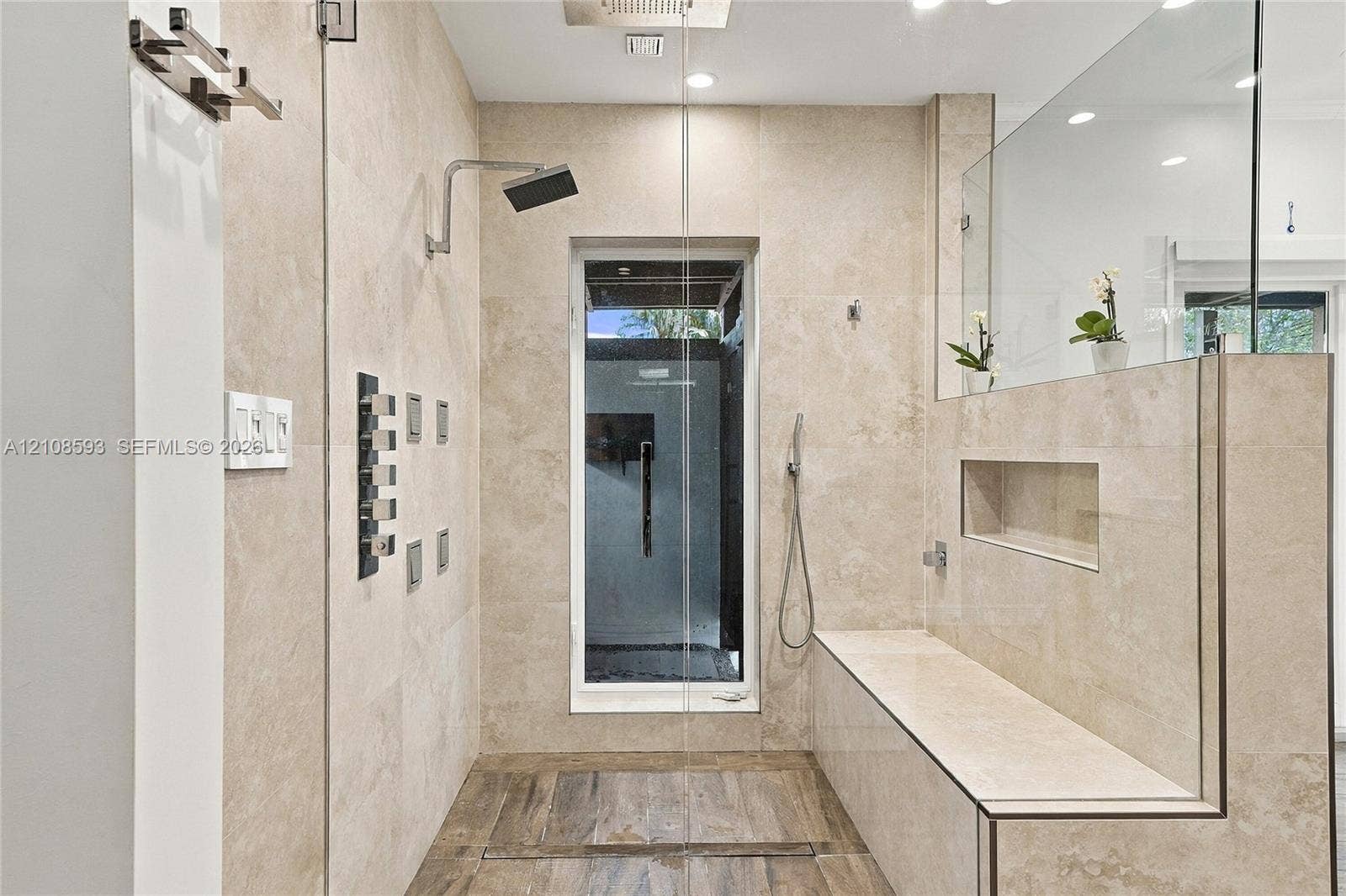 8230 SW 151st Street, Palmetto Bay, FL 33158 Walk-in shower featuring large-format neutral stone wall tile, multi-head shower system with a rain shower head, built-in stone bench with integrated recessed shelving, and wood-finish flooring