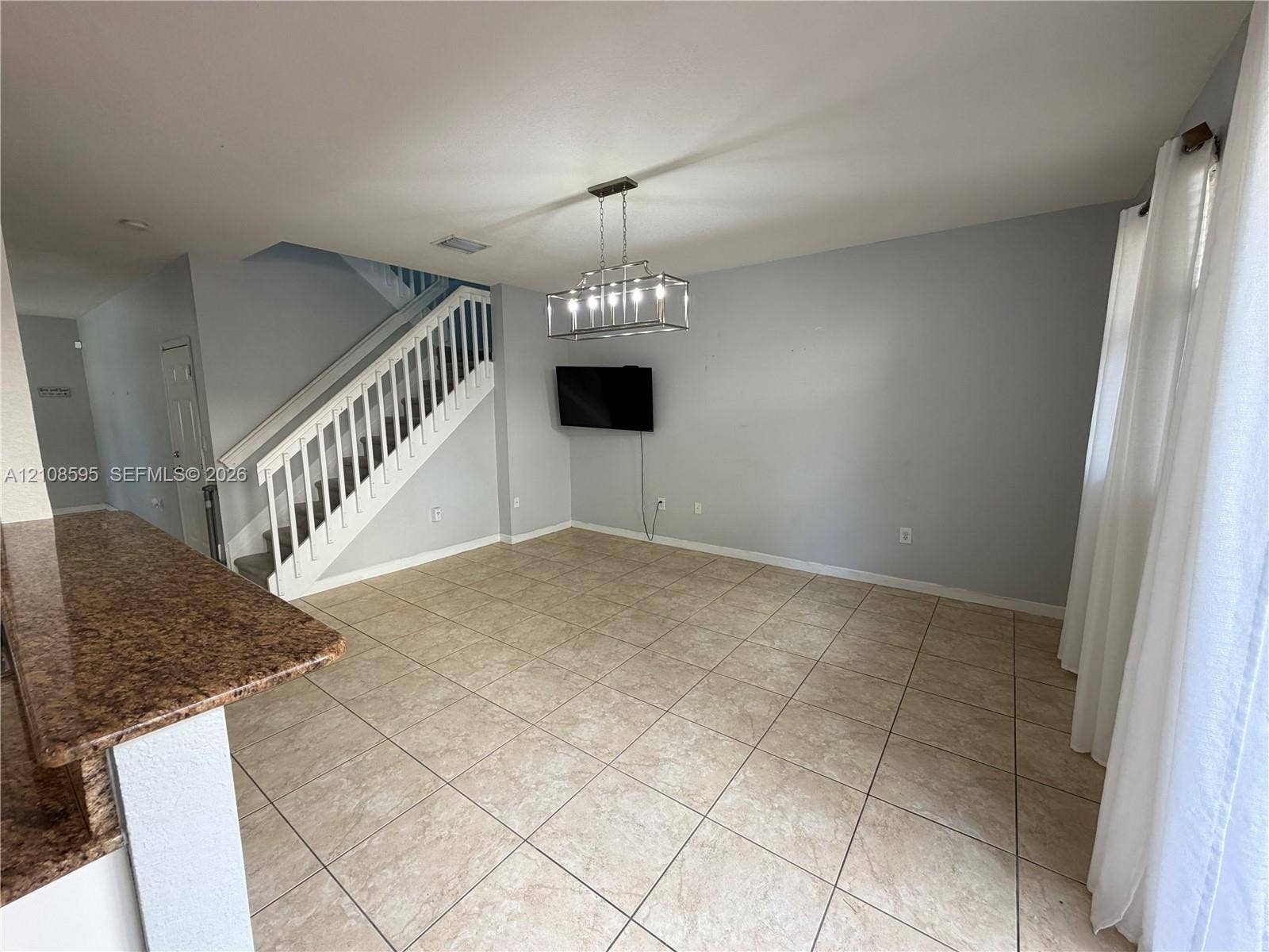 12989 SW 135th Terrace, #0, Miami, Miami, FL 33186 Open-concept interior featuring large-format tile flooring, stone-surfaced peninsula countertop, staircase with white balusters, and a rectangular multi-light chandelier