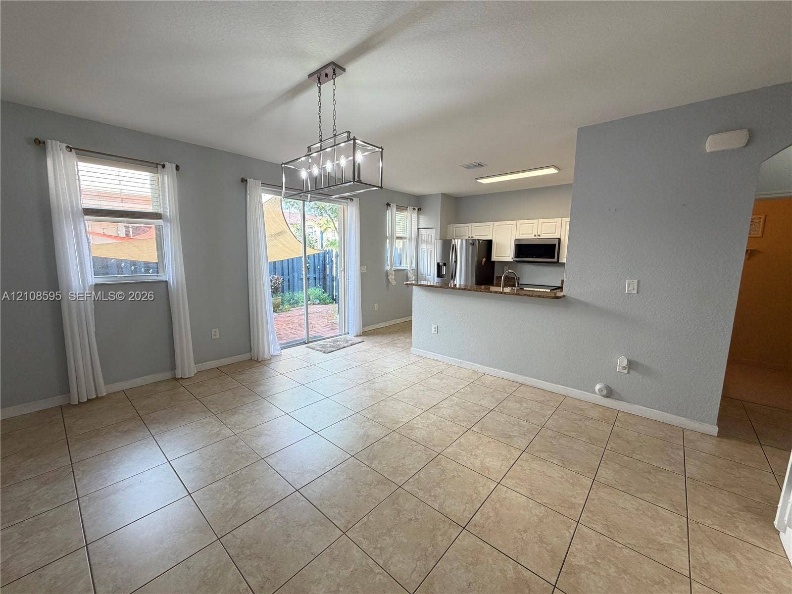 12989 SW 135th Terrace, #0, Miami, Miami, FL 33186 Open-concept interior featuring large-format ceramic tile flooring, a geometric metal chandelier, white kitchen cabinetry, and a sliding glass door providing exterior access