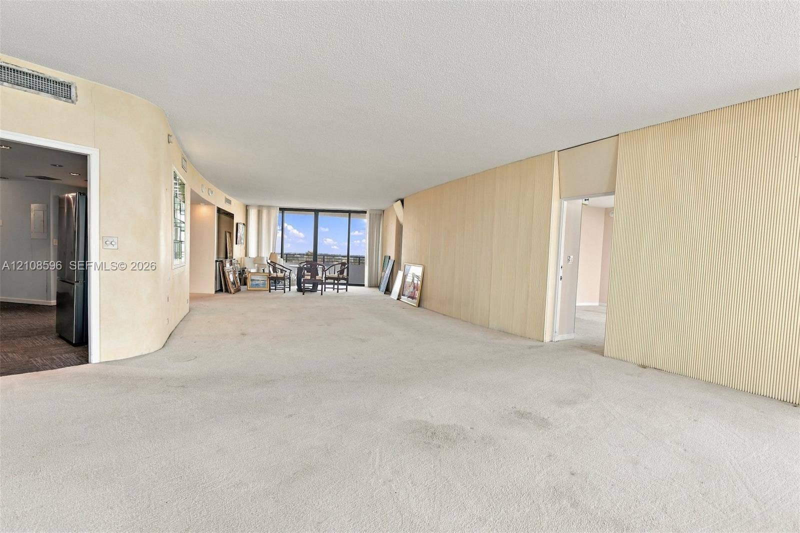 Open-concept floor plan featuring expansive light-toned wall paneling, curved wall architecture, floor-to-ceiling glass sliding doors, and neutral wall-to-wall carpeting