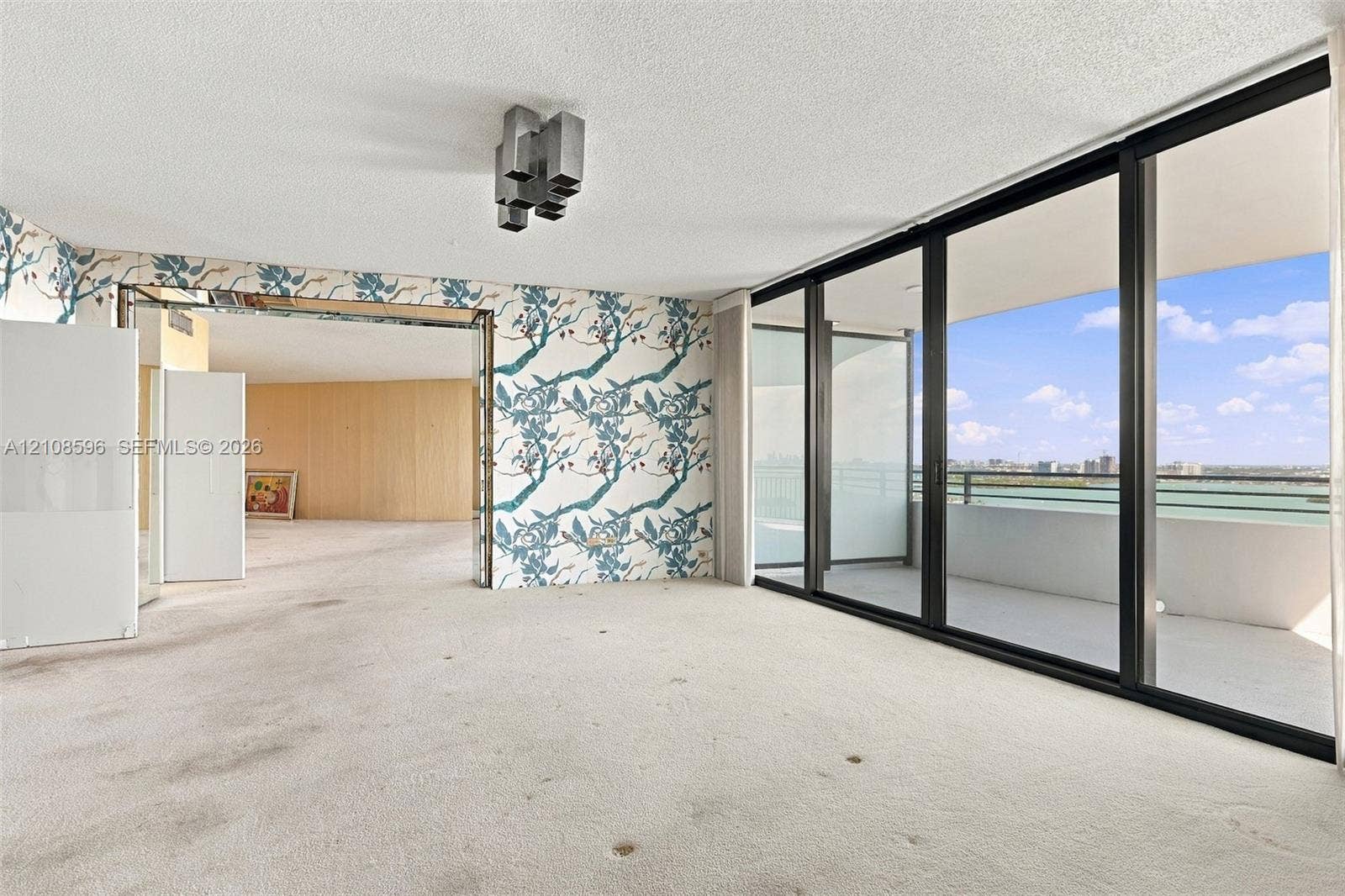 Floor-to-ceiling glass sliding doors opening to an expansive private balcony, featuring a multi-panel black frame and geometric ceiling-mounted lighting