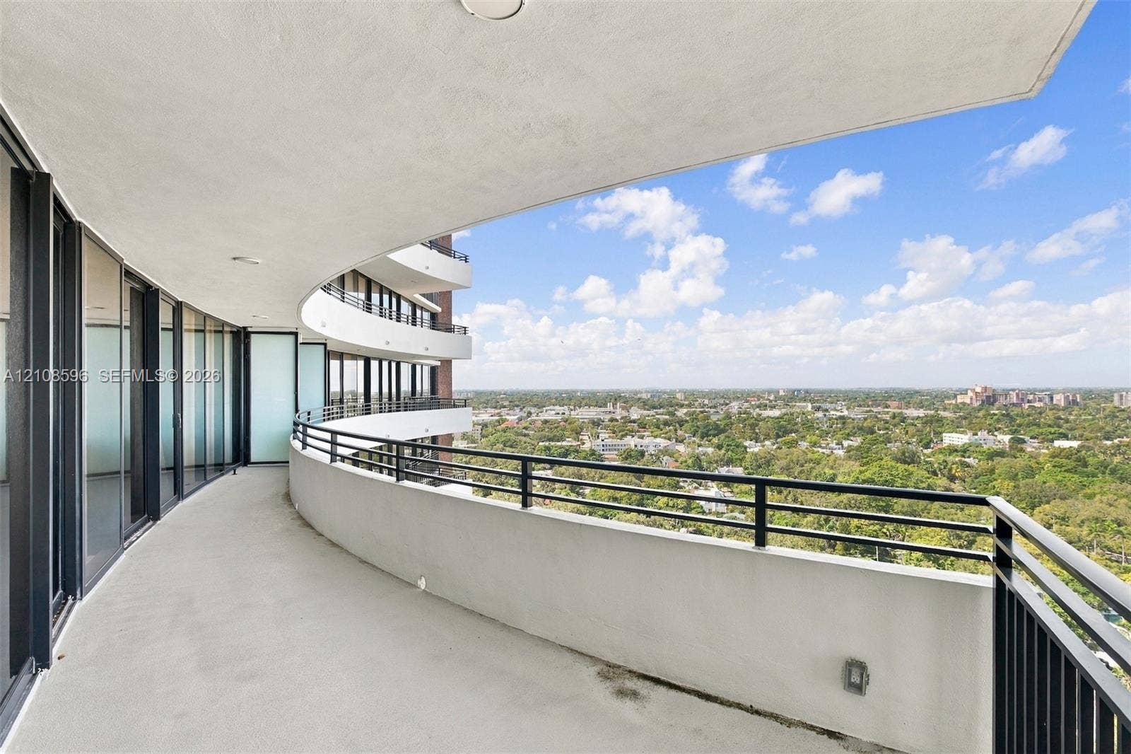 Curved exterior balcony featuring concrete flooring, a solid half-wall, and black metal railings with panoramic outdoor surroundings
