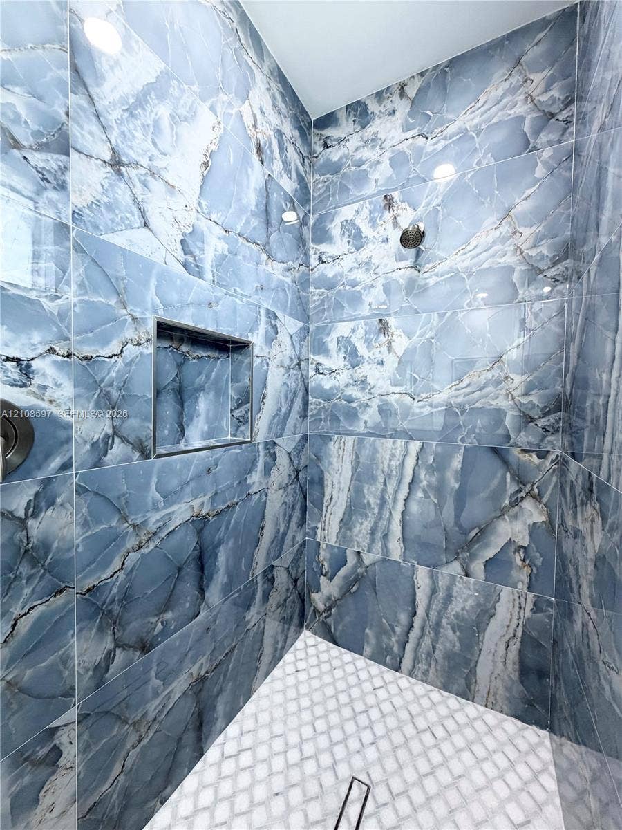 808 Congress Avenue, Lehigh Acres, FL 33972 Shower stall featuring full-height blue-toned stone-patterned wall tiles, integrated recessed shelving, white geometric mosaic floor tiling, and a metallic shower fixture