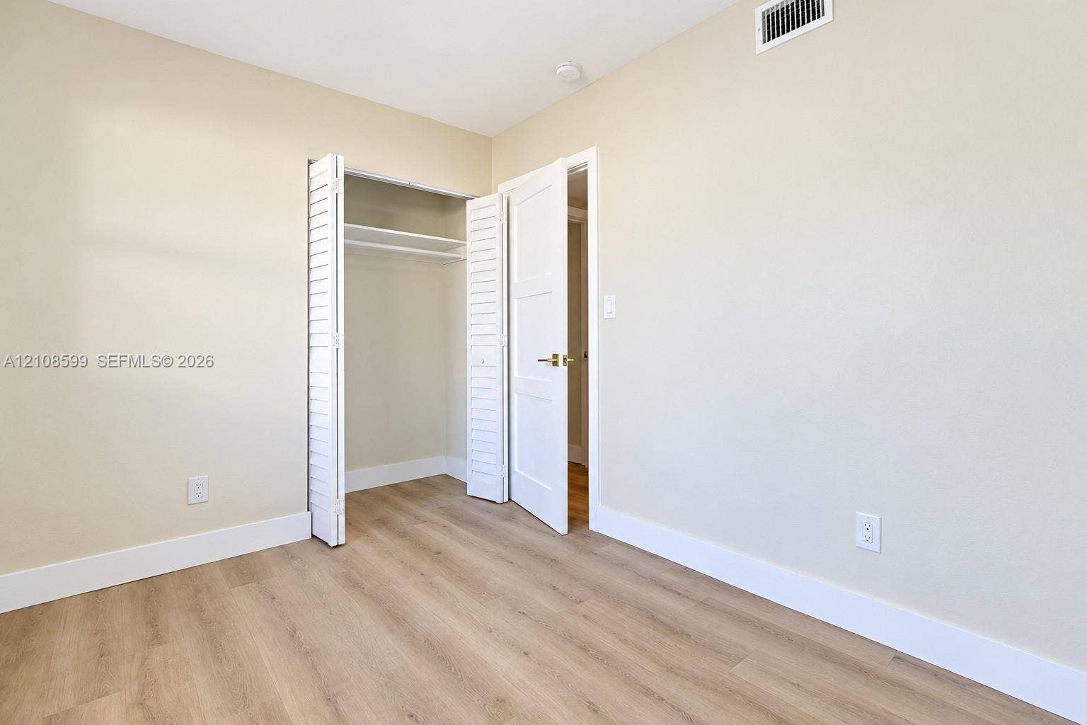 Wood-finish flooring, neutral wall tones, white louvered closet doors with interior shelving, and a white interior door featuring brass-tone hardware