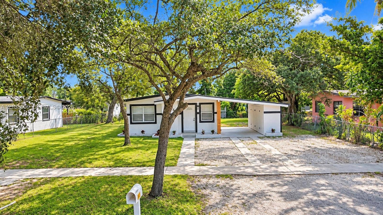 Single-story exterior featuring a flat roofline, attached carport, gravel driveway, and a large front lawn with mature trees