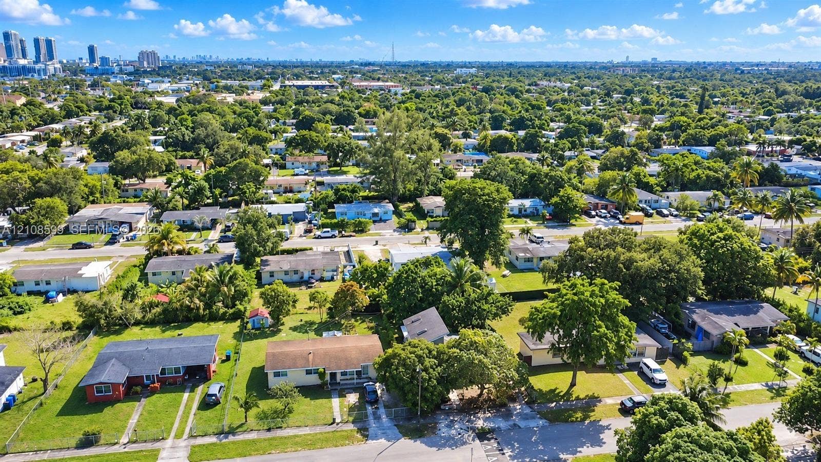 Aerial perspective of a suburban neighborhood featuring tree-lined streets, residential lots with manicured lawns, and a distant city skyline