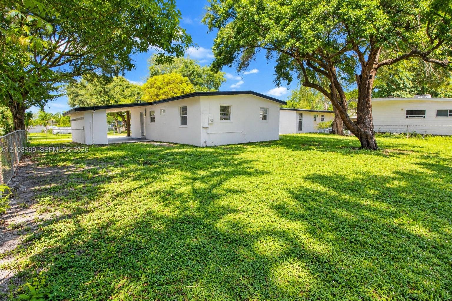 Exterior structure featuring a white stucco finish, covered side porch, mature trees, and chain-link perimeter fencing