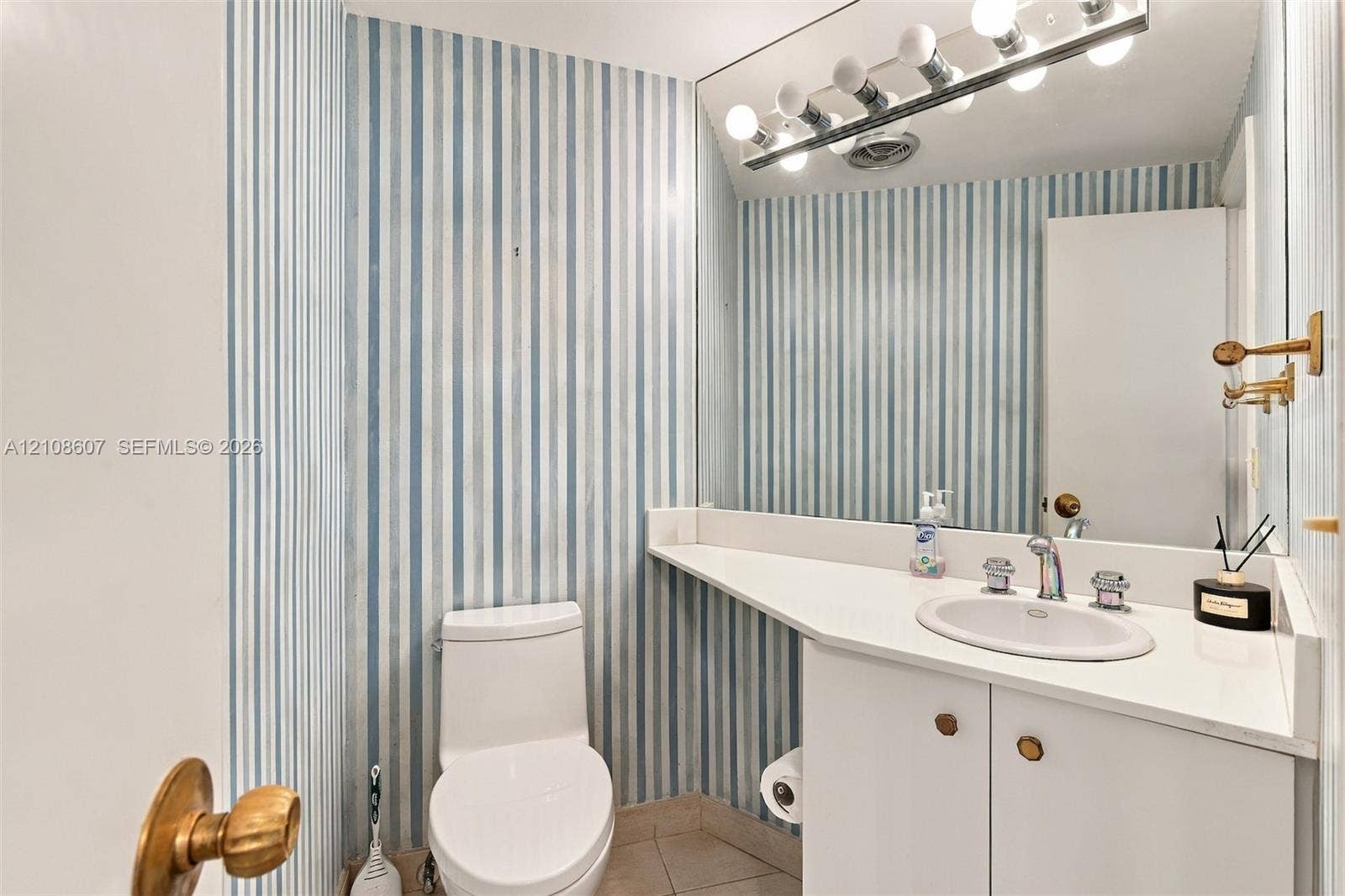 Bathroom featuring a white cabinet with integrated countertop and recessed sink, wall-spanning mirror, multi-bulb vanity light fixture, and vertical-striped wall covering