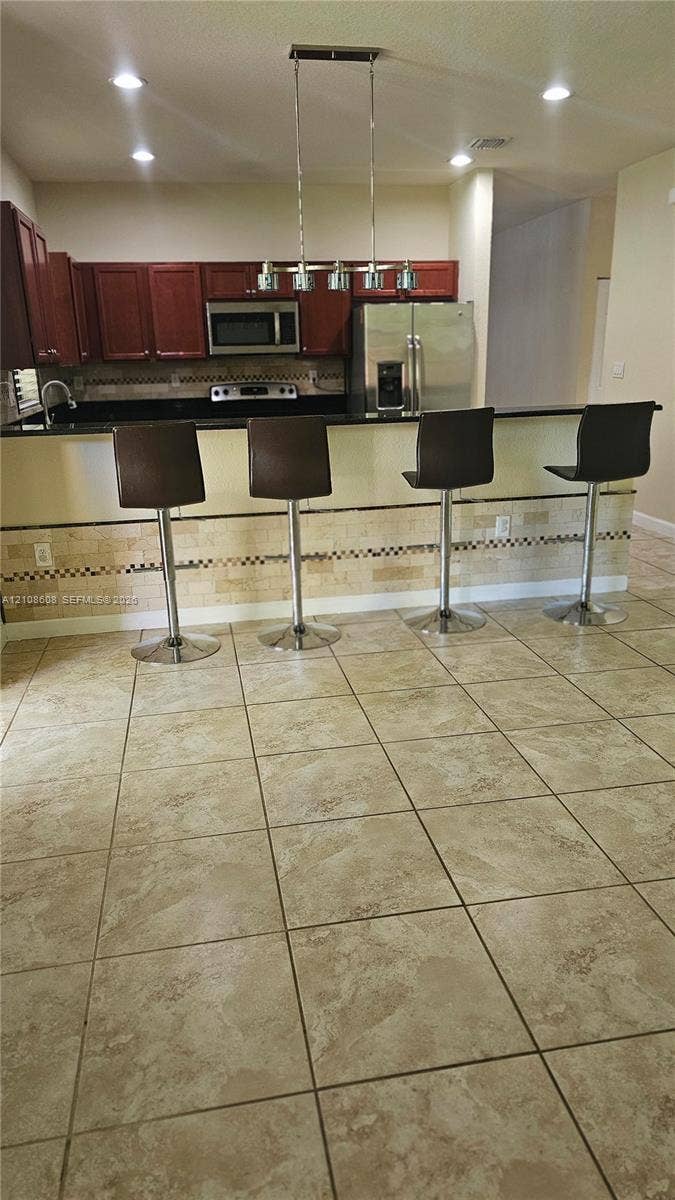 24041 SW 114th Place, Homestead, FL 33032 Kitchen featuring a peninsula island, stainless steel appliances, tile flooring, stone-tiled backsplash, and overhead pendant lighting