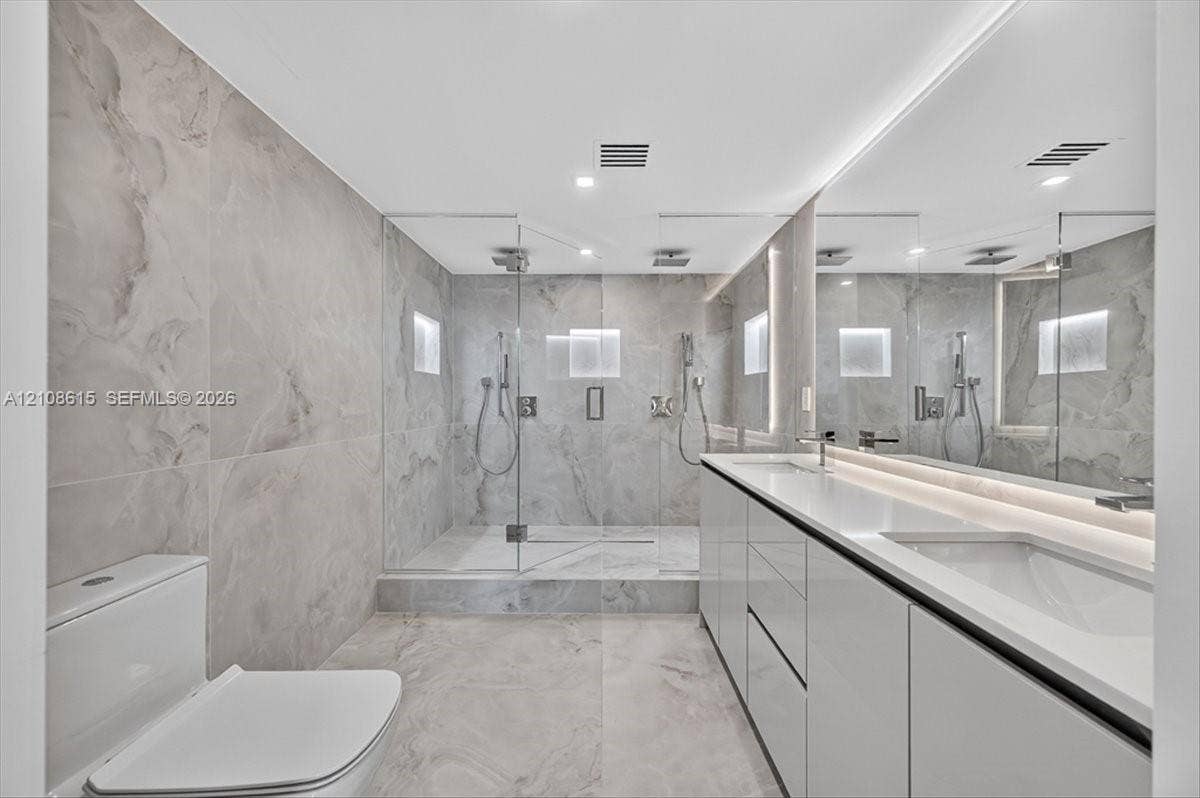9455 Collins Avenue, #1201, Surfside, FL 33154 Dual-sink vanity with high-gloss cabinetry and solid surface countertop, walk-in glass-enclosed shower featuring dual rainfall showerheads and recessed wall niches, and floor-to-ceiling large-format tile surround