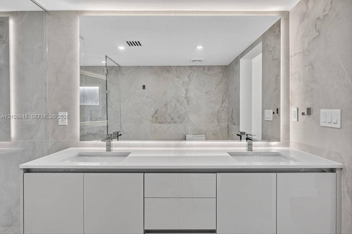 9455 Collins Avenue, #1201, Surfside, FL 33154 Modern dual-sink vanity featuring a solid-surface countertop, high-gloss cabinetry, integrated LED-backlit mirror, and polished metal fixtures against full-height stone-finish wall tile