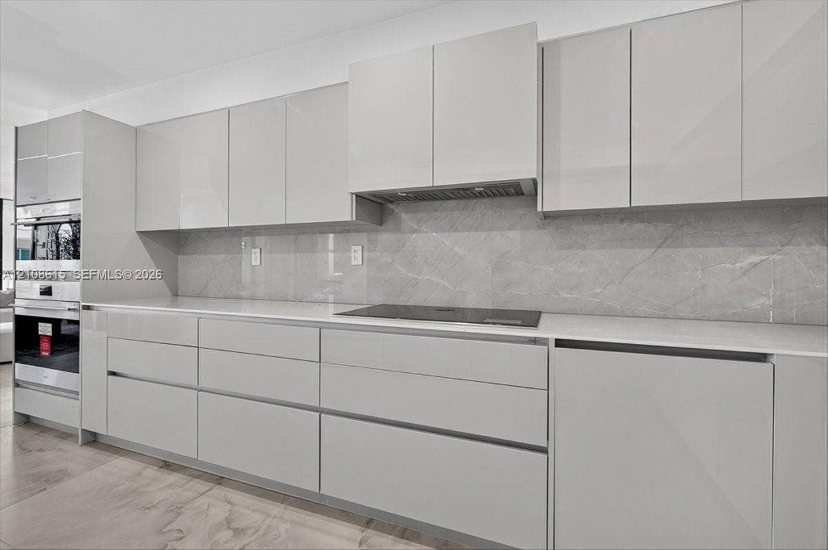 9455 Collins Avenue, #1201, Surfside, FL 33154 Sleek kitchen featuring high-gloss cabinetry, expansive stone-finish backsplash, built-in wall oven, flush-mount cooktop, and light-toned countertops