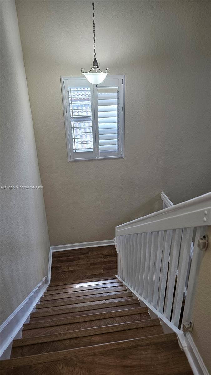 Staircase featuring wood-finish treads, a white balustrade, a window with interior shutters, and a pendant light fixture