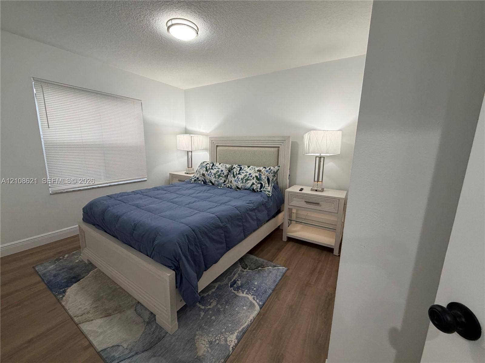 Neutral wall tones, wood-finish flooring, and flush-mount ceiling lighting, complemented by a window with horizontal blinds