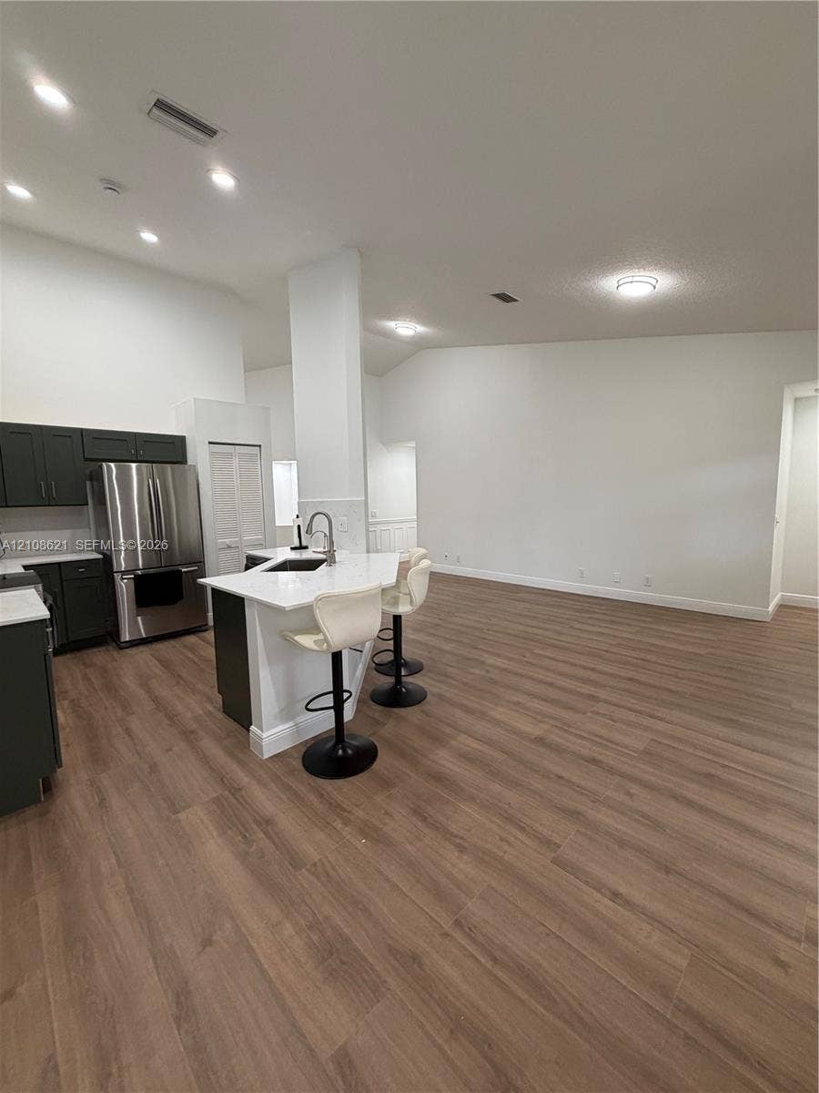 Kitchen featuring wood-finish flooring, dark cabinetry, stainless steel refrigerator, white countertops, and a central island with a built-in sink and faucet