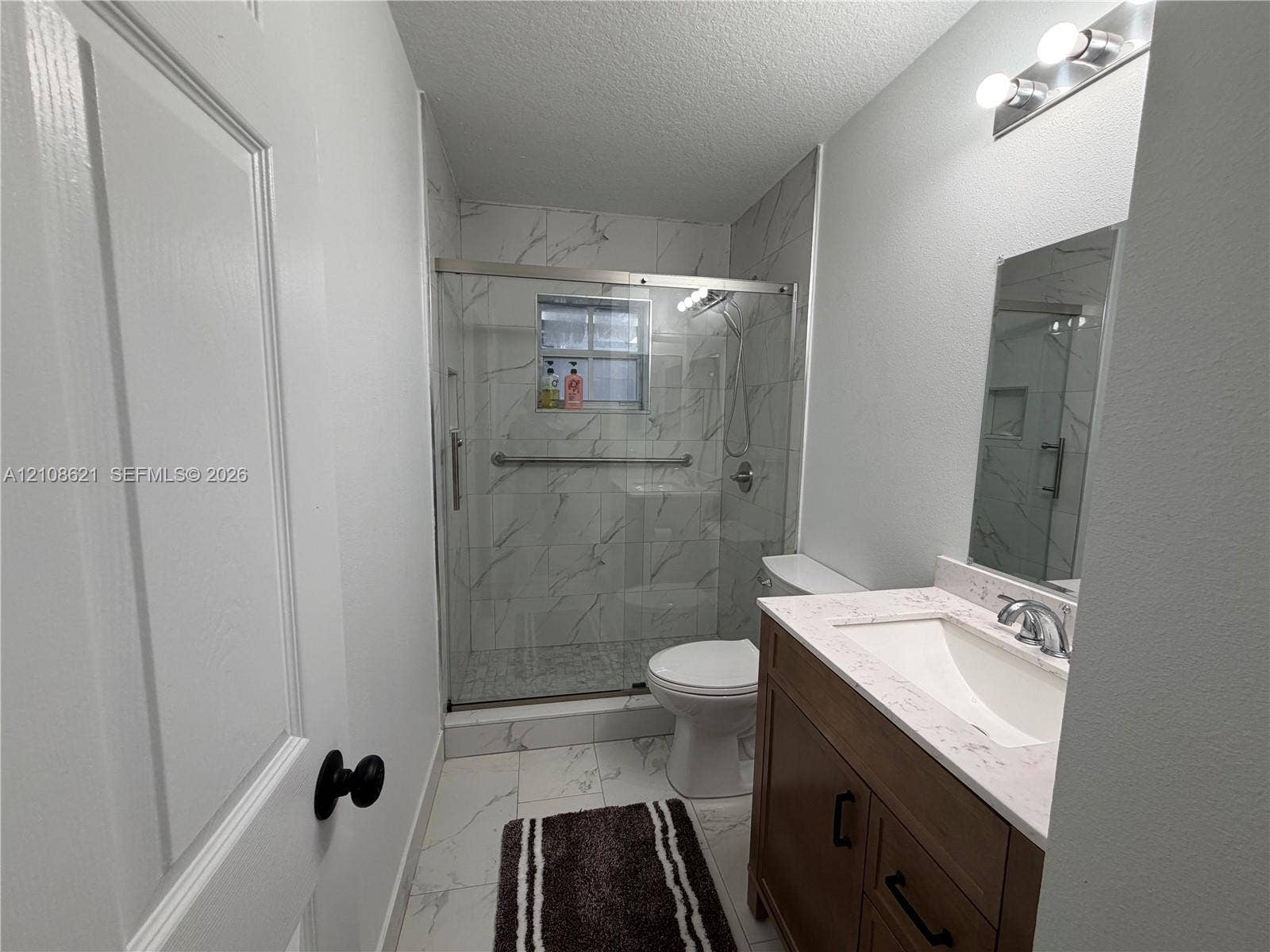 Bathroom featuring a vanity with a light-toned countertop, integrated basin, and matte black hardware, paired with a glass-enclosed shower with stone-patterned wall tile and matching floor tile