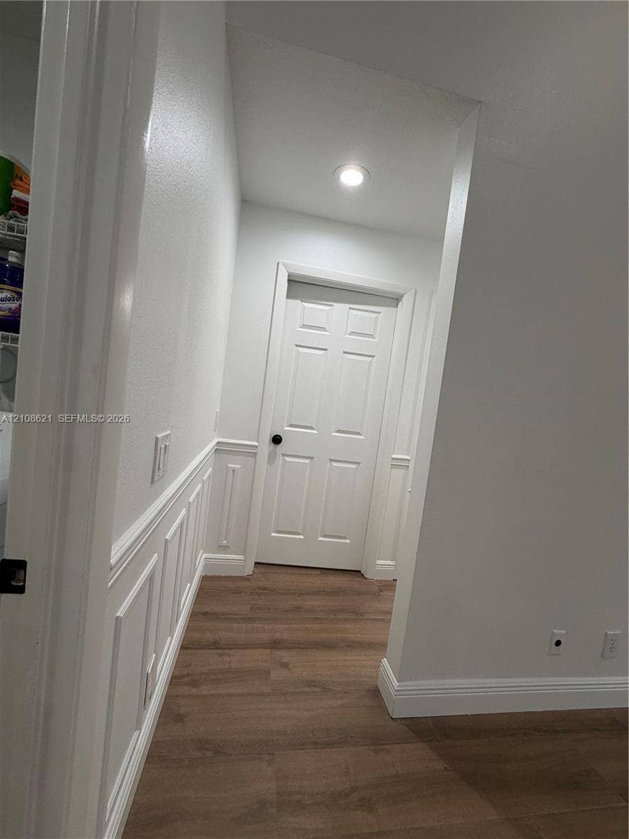 Interior hallway featuring wood-finish flooring, white wainscoting, a six-panel door with matte black hardware, and recessed ceiling lighting