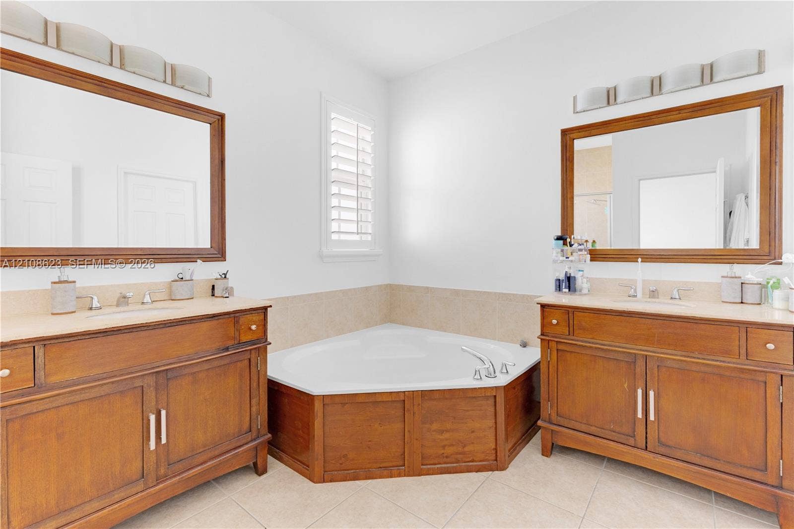 Dual wood-finish vanities with light-toned countertops, corner soaking tub with wood-finish surround, two rectangular wall-mounted mirrors, and tiled flooring