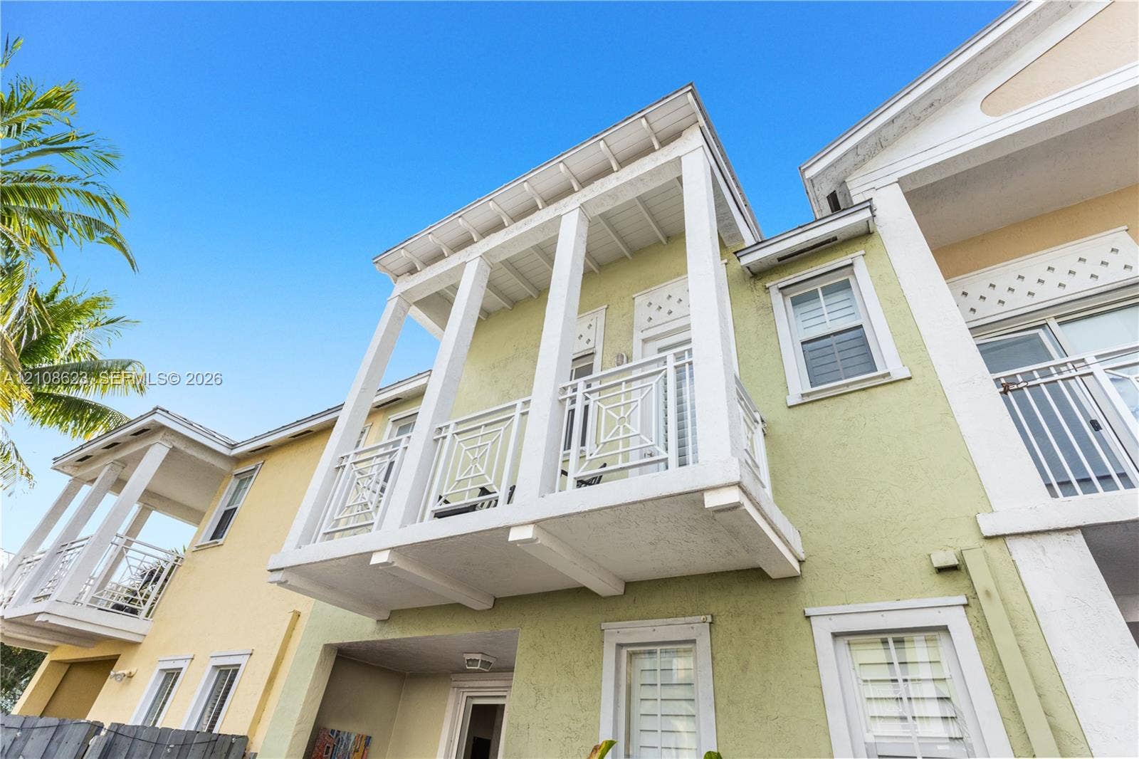 Multi-level exterior featuring light-toned stucco, a covered upper-level balcony with geometric railing, and white-framed windows with plantation shutters