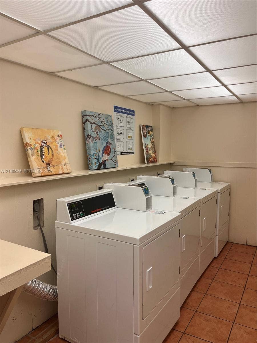 3650 Inverrary Drive, #1Q, Lauderhill, FL 33319 Laundry facility featuring multiple white front-loading appliances, a built-in wall shelf, a drop ceiling with integrated lighting, and tile flooring