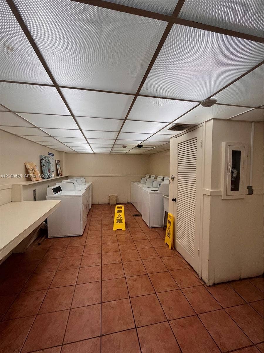 3650 Inverrary Drive, #1Q, Lauderhill, FL 33319 Laundry facility featuring ceramic tile flooring, a suspended grid ceiling, built-in folding counter, and multiple washing and drying units