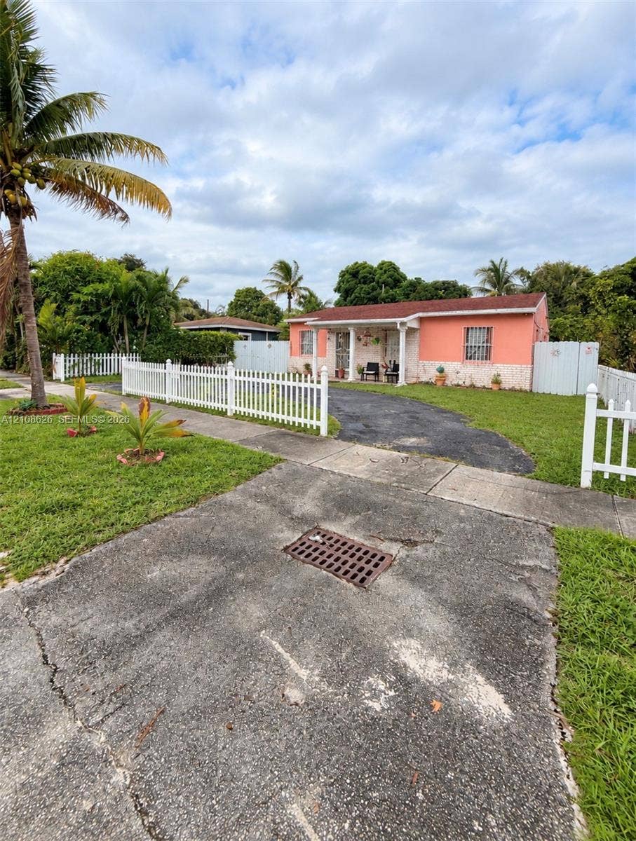 971 NE 156th Terrace, North Miami Beach, FL 33162 Single-story exterior featuring a vibrant stucco finish, stone-veneer foundation, covered front porch with columns, white picket fencing, and paved driveway
